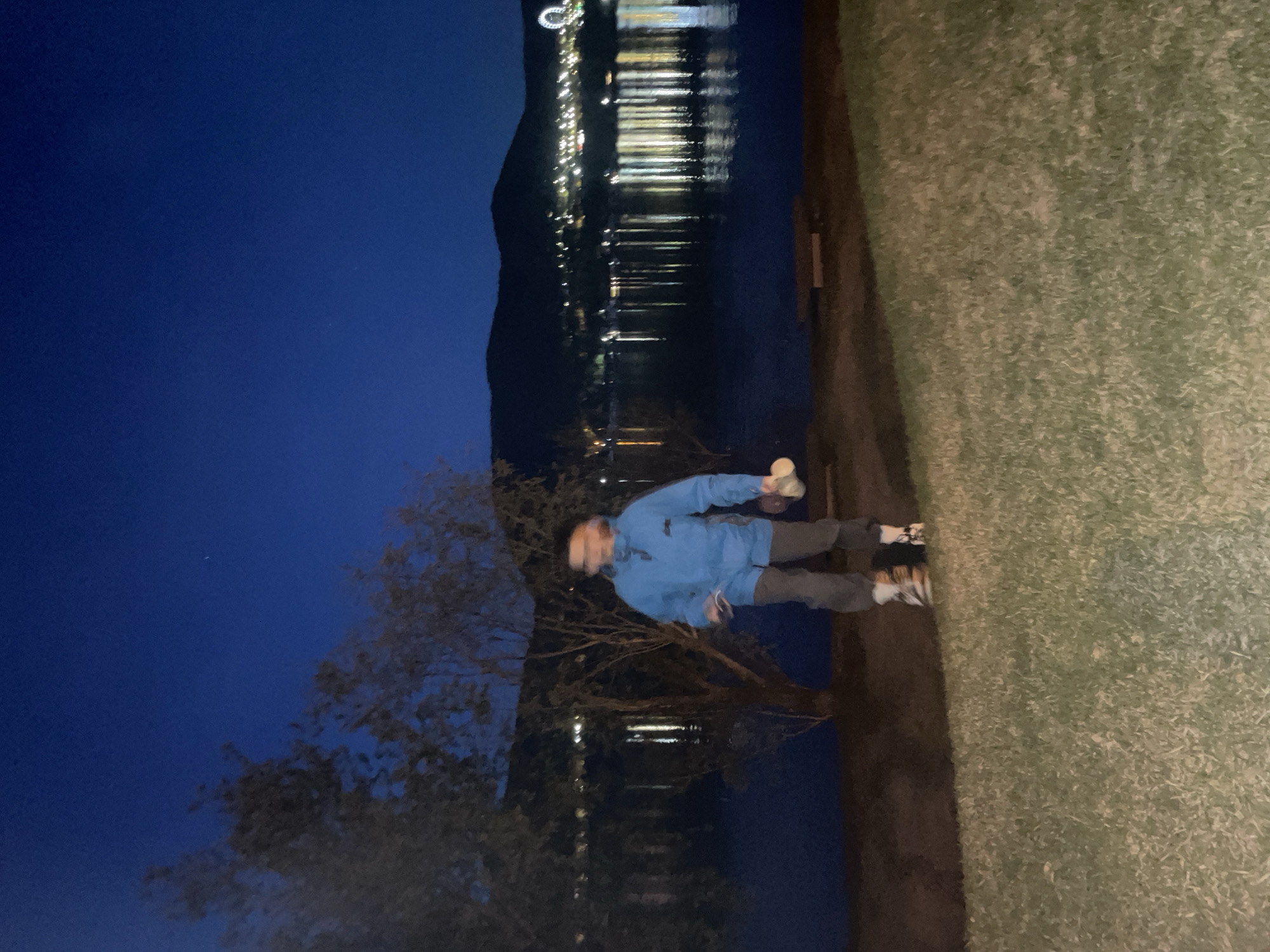 A nighttime photograph captures a person standing next to a body of water. The person is wearing a blue jacket and grey pants. In their hand, they appear to be holding a white object. Behind the person is a tree, and beyond the water, there are city lights reflecting off the surface. The other side of the frame shows grass. The sky is a dark blue hue. The location seems to be in the city of Krasnoyarsk, Russia.