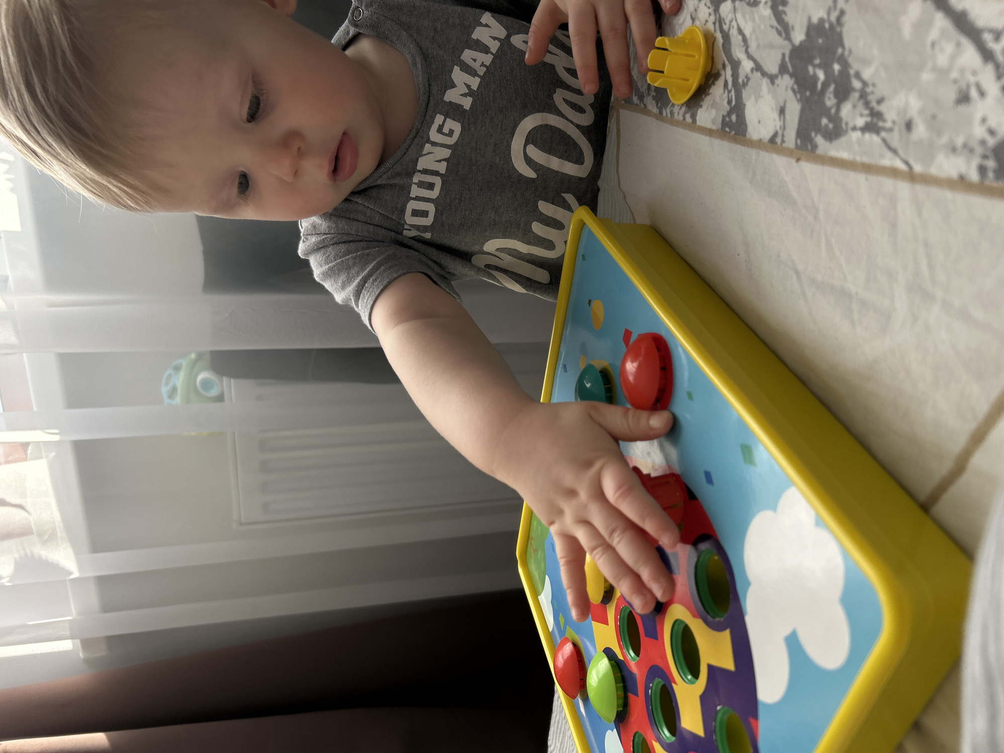 A young child with fair skin and light blonde hair, wearing a grey short-sleeved t-shirt with "YOUNG MAN My Dad" printed on it, is seated at a table. The child's right hand is extended over a brightly colored plastic pop-up toy, actively interacting with or about to press a red domed button. The toy features a yellow base and a blue surface with white clouds, circular holes revealing colorful internal patterns, and several red, green, and blue domed buttons, some of which are depressed. A small, separate yellow plastic piece rests on the tablecloth to the child's right. The table is covered by a light-colored tablecloth, split by a seam into a plain section and a section with a mottled grey and white pattern. In the background, a window draped with sheer white curtains is visible, admitting natural light. A white heating radiator is installed beneath the window, and a dark brown curtain panel is present on the far left. The setting is an indoor residential environment, consistent with a home in Chaikovsky, Russia.