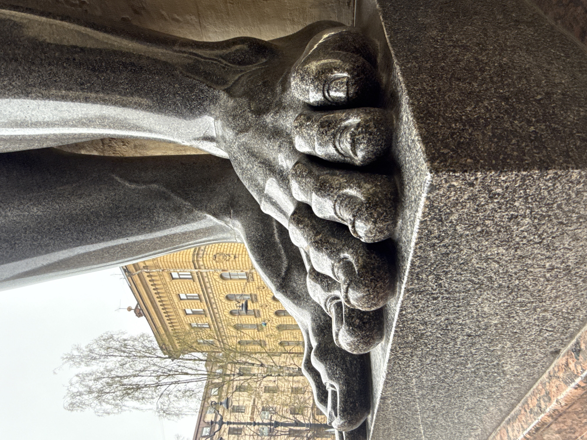 The image is a close-up of the foot of an Atlant statue supporting a building's facade. The statue is made of granite. In the background, a light-colored building with several windows and ornate decoration is visible. A leafless tree is partially obscuring the building. The statue is located in Saint Petersburg, Russia.