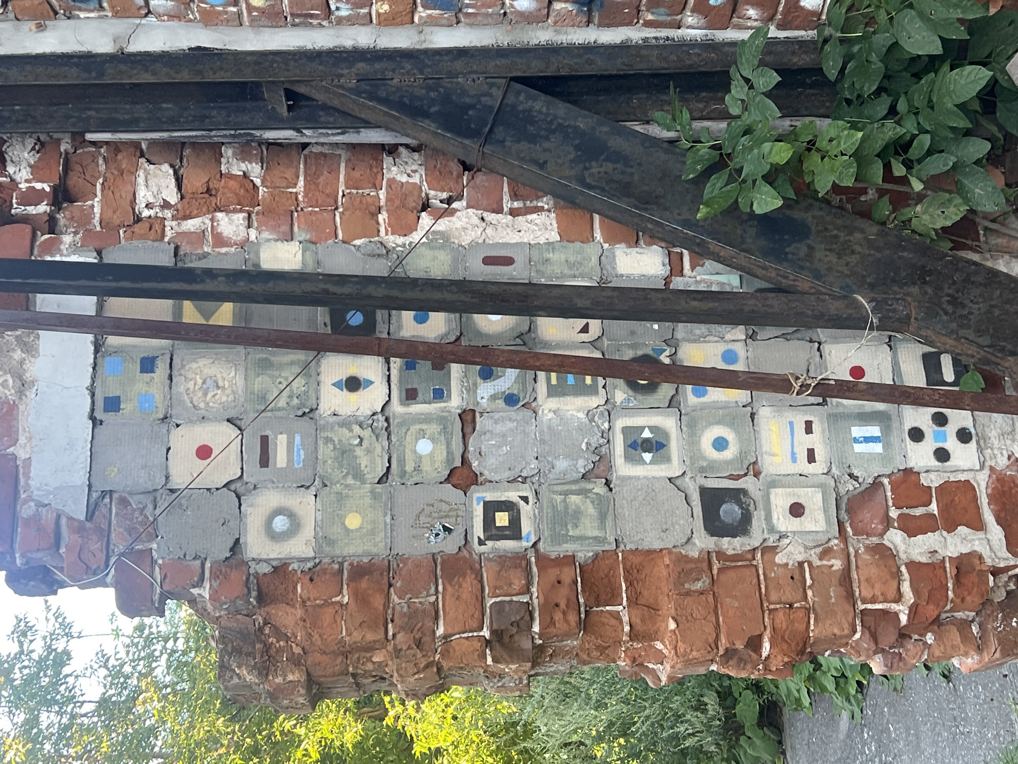 This is a close-up, eye-level outdoor shot of a damaged brick wall adorned with a mosaic of square tiles, located in Nizhny Novgorod, Russia. The wall is partially crumbling, revealing older red brickwork and newer grey concrete infill. The mosaic tiles, each approximately 4x4 inches, are arranged in a grid and feature simple geometric designs and solid colors, including red circles, blue squares, yellow circles, and brown and blue rectangles. Some tiles are chipped or faded, with a few missing entirely.
Behind the tiled section, two vertical, rusted metal pipes run parallel, and a larger, angled rusted metal beam is attached to the wall. Green foliage from a nearby plant is growing up around the metal structures and against the wall. In the background, on the left side of the frame, lush green trees and foliage are visible under a bright sky. The ground in front of the wall appears to be a paved surface, possibly a sidewalk or road.