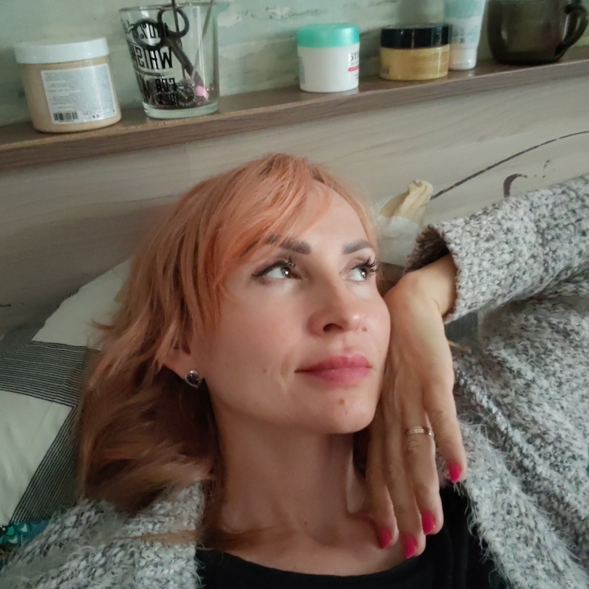 An indoor medium close-up shows a woman, likely in her 30s or 40s, with light reddish-blonde hair and bangs, lying down and looking upwards with a contemplative expression. She has visible makeup, including dark eyeliner, mascara, pink lipstick, and filled eyebrows. She wears a dark stud earring on her left ear and a thin silver-colored ring on her right ring finger. Her fingernails are painted bright pink. She is dressed in a light grey, fuzzy, textured sweater or cardigan.
Behind her, a wooden shelf holds several items: a beige jar with a white lid and a label (partially visible text and a barcode), a clear glass containing scissors (with reversed text "SO" and "BIHW" visible), a white jar with a light blue/turquoise lid (with "ST" visible), a yellow jar with a black lid, a white tube, and a dark brown/black mug. The wall behind the shelf is a light green/teal color. A striped pillow (white with dark stripes) is visible to her left, suggesting a bedroom setting. The location context provided is Chaikovsky, Russia. The image captures a relaxed, casual moment.