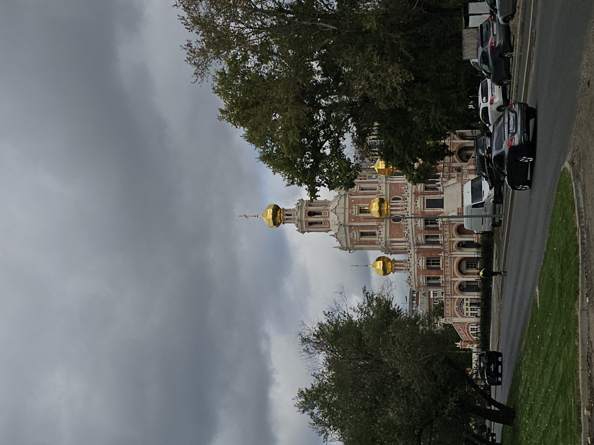 The image shows a street scene in Moscow, Russia, with the Church of the Intercession at Fili in the background. The church is a prominent pink and white building with multiple golden domes. In the foreground, cars are driving on a road, and there is a grassy area to the left. A person in a yellow jacket is walking on the side of the road. The sky is overcast and cloudy.