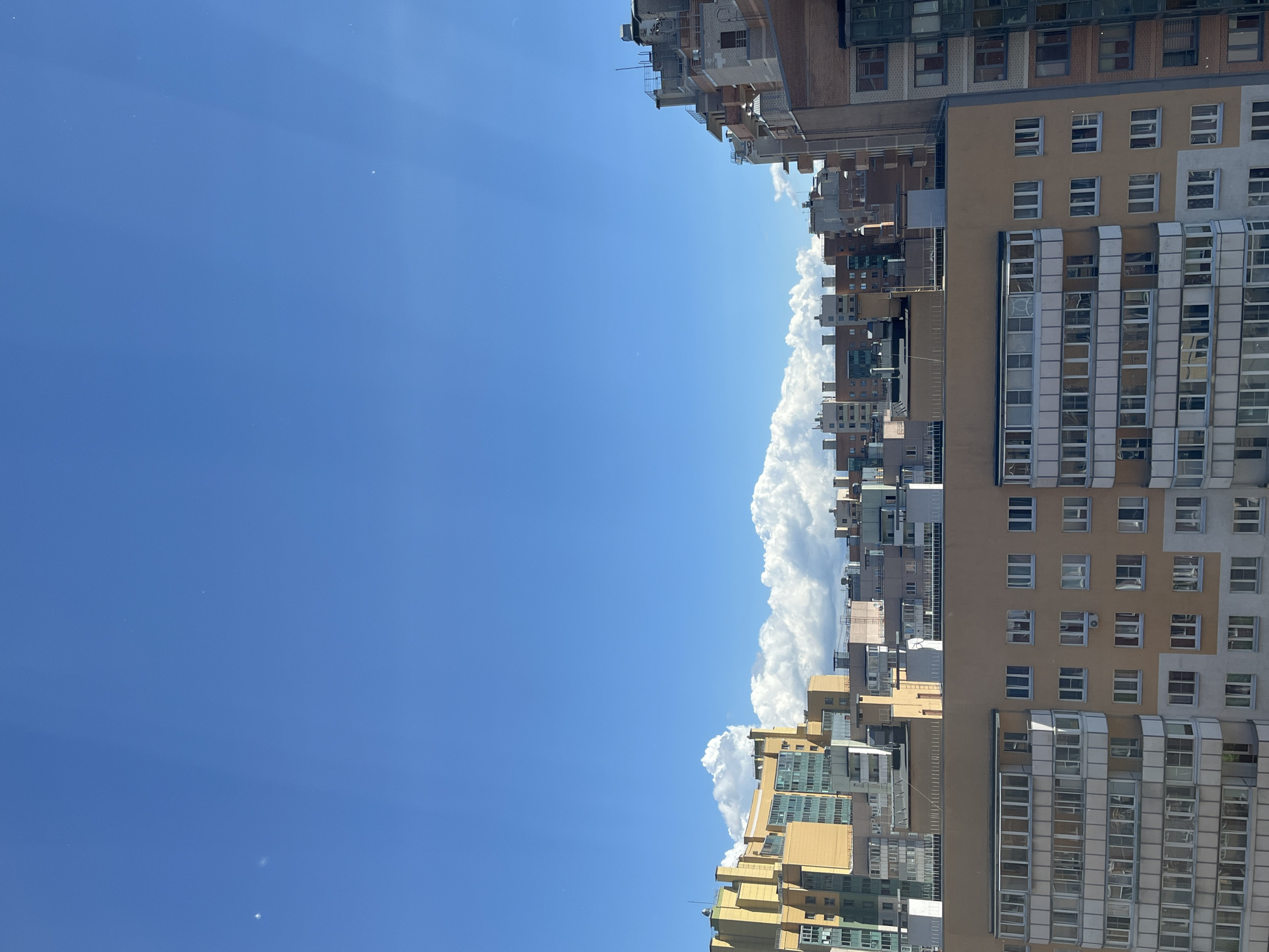The image captures a view of a cityscape under a clear blue sky with scattered white clouds. The foreground features a multi-story apartment building with beige and white facades and numerous windows. Behind this building, a collection of other apartment buildings of varying heights and architectural styles are visible. Some buildings are brightly colored in yellow and green, while others have more muted brown and gray tones. The buildings are densely packed, indicating an urban environment. The sky is a vibrant blue, with sunlight casting subtle rays that are visible as faint streaks. There are no people or vehicles clearly discernible in the image. The scene appears to be a typical residential area in a Russian city.