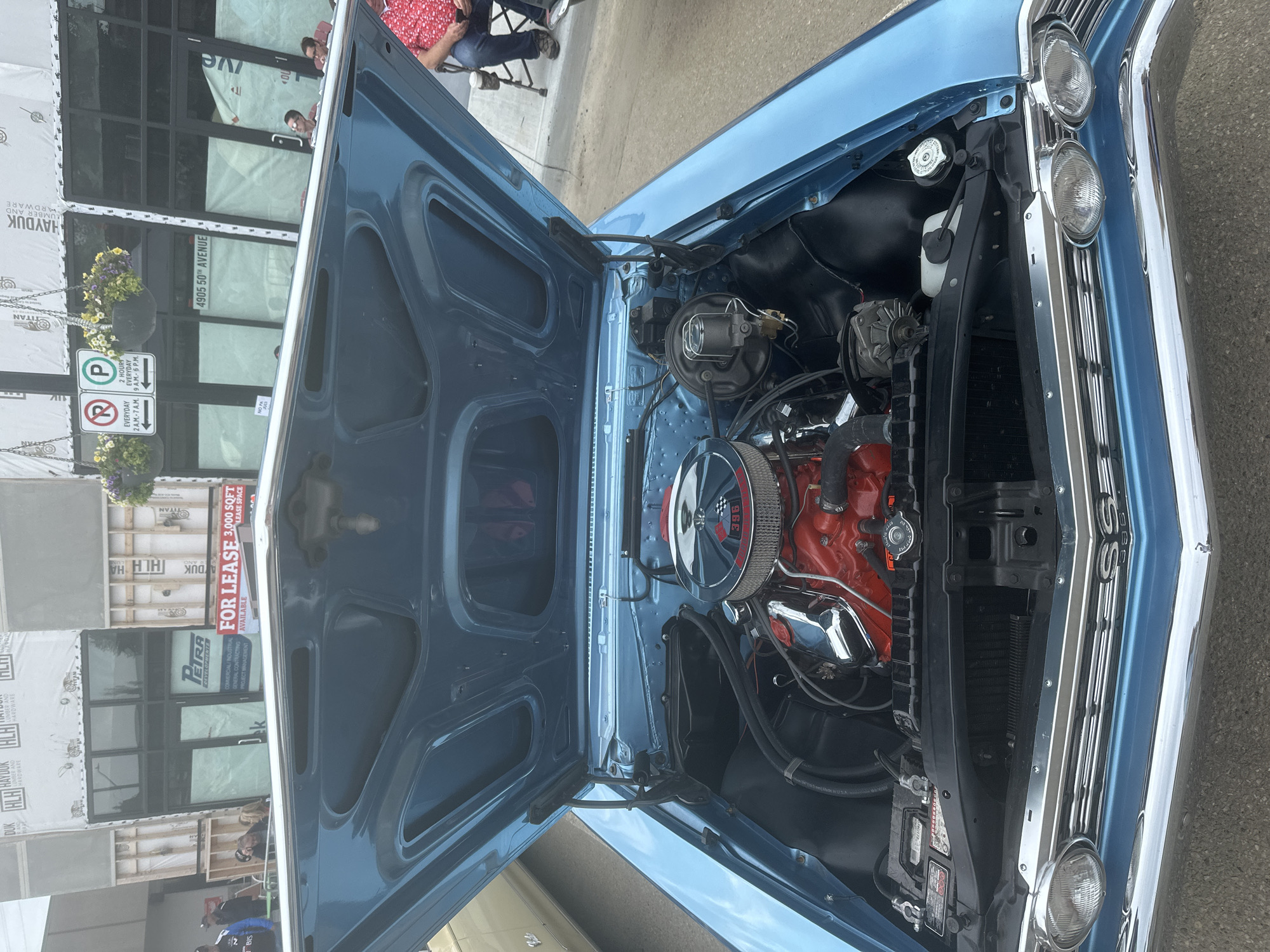 The image shows the open hood of a light blue classic car, revealing its engine. The car is parked on a street in front of a building undergoing construction. In the background, there is a sign that reads "FOR LEASE 3,000 SQFT LEASE SPACE," indicating a commercial property. Another sign on the building shows the address "4905 50th Avenue." This address corresponds to a location in the City of Leduc, Alberta, Canada. There are several people visible in the background, some standing and others sitting in chairs, suggesting an outdoor event or gathering. The overall scene depicts a street view with a focus on the vintage automobile and its engine.