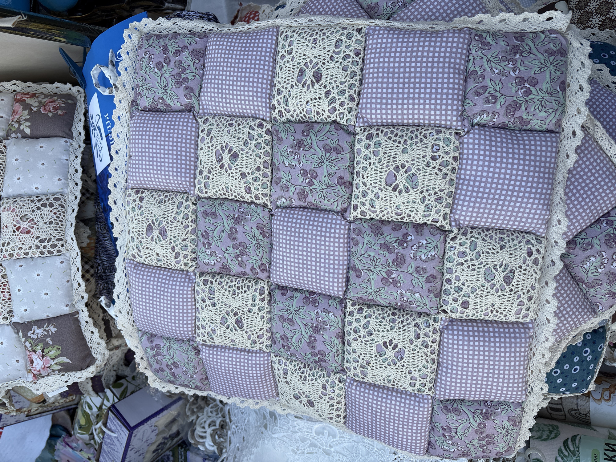 The image shows a close-up view of a handmade quilted cushion or mat. The cushion is made of small square patches of fabric. These patches are in various patterns, including a lilac gingham check, a lilac fabric with a floral pattern of small purple flowers and green leaves, and square patches of cream-colored crochet lace. The entire cushion is bordered with a decorative cream lace trim. The cushion appears to be placed on top of other items, possibly at a market stall, as suggested by the out-of-focus background which includes other textiles and boxes. No people are visible in the image. The lighting is natural, suggesting an outdoor or well-lit indoor setting. The specific location within Vitsebsk, Belarus cannot be determined from the visual information provided.