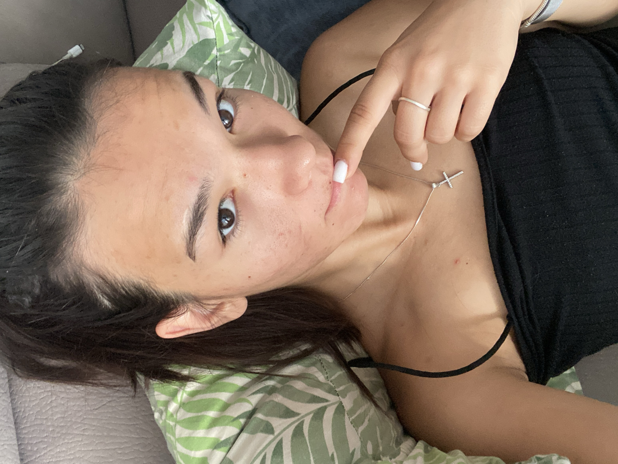 A young woman with dark hair pulled back lies on a couch, resting her head on a pillow with a green and white leaf pattern. She is wearing a black strappy top and a silver necklace with a cross pendant. Her right hand is brought to her mouth, with white-painted fingernails and a silver ring on her index finger. A small red mark is visible on her upper chest. The background suggests an indoor setting. The image does not provide any specific information about the location in Plovdiv, Bulgaria.
