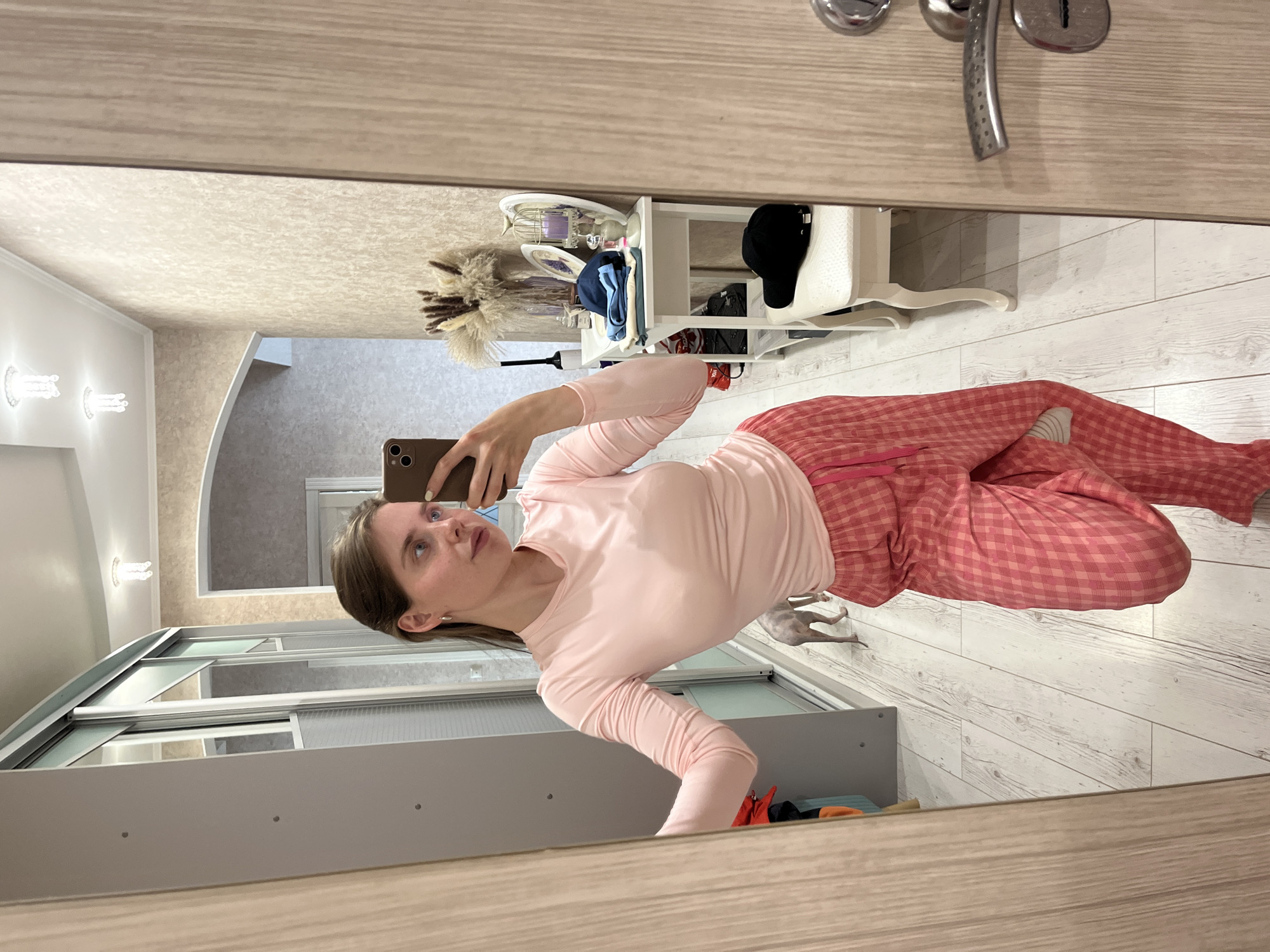 A woman is taking a selfie in a mirror in a room in Chaikovsky, Russia. She is wearing pink pajamas and holding a smartphone. The room has a wooden floor and a white wall. There is a white table with a hat, dishes, and decorations on the background. The image appears to be taken in a residential setting.