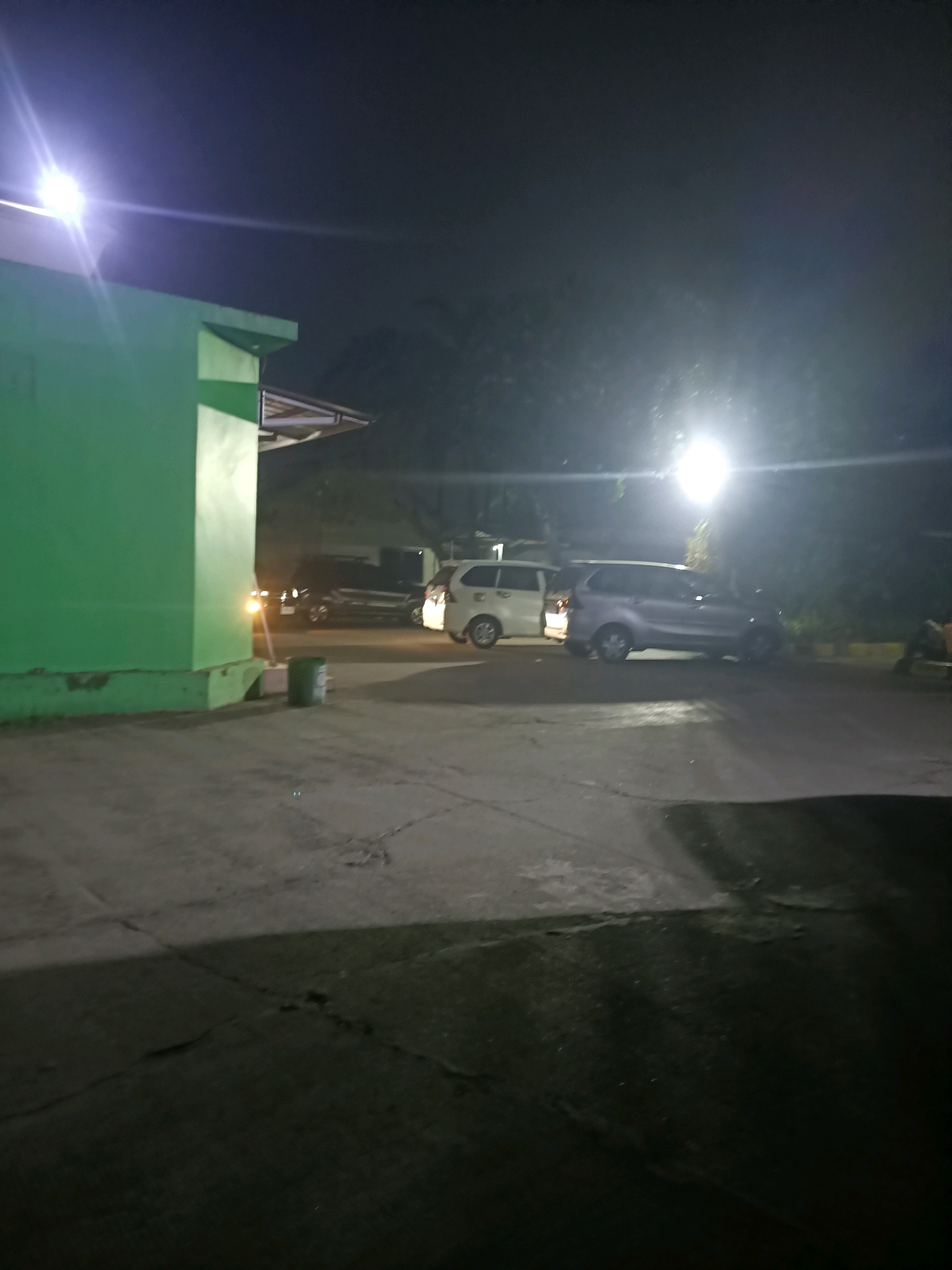 The image depicts an outdoor scene at night, likely a parking area or a service road, in Karawang, Indonesia. A bright light fixture illuminates the scene from the upper left. A prominent green building, with some architectural features like a protruding roof overhang, occupies the left side of the frame. In the background, two vehicles are visible: a silver minivan and a white minivan. A dark-colored car is parked behind the green building. The ground surface appears to be paved, with some cracks and debris. A green cylindrical trash bin is situated near the base of the green building. The overall impression is of a utilitarian or industrial area at night, with artificial lighting casting a glow on the surroundings.
