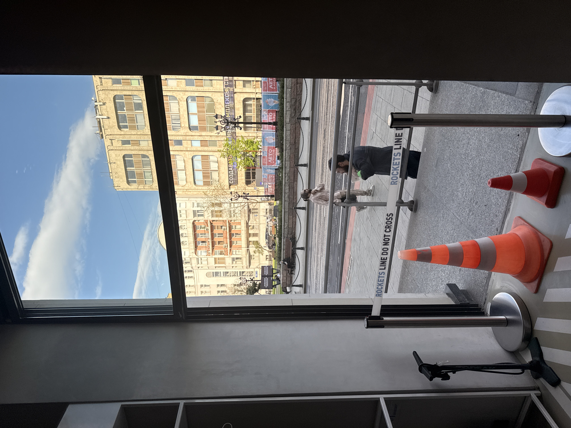 The image shows a view through a window, possibly from indoors. Outside, a city street scene unfolds, potentially in Moscow, Russia, given the partially visible Cyrillic script on banners. Pedestrians are present, including a person bending over and another walking. The street has what appears to be trolley tracks. Sidewalk barricades, a "ROCKETS LINE DO NOT CROSS" sign, and traffic cones are noticeable. The architecture across the street includes tall buildings with classical design elements. The sky visible above the buildings is blue with scattered clouds. Indoors, the window frame is visible, along with sections of a shelf and what appears to be a bicycle pump.