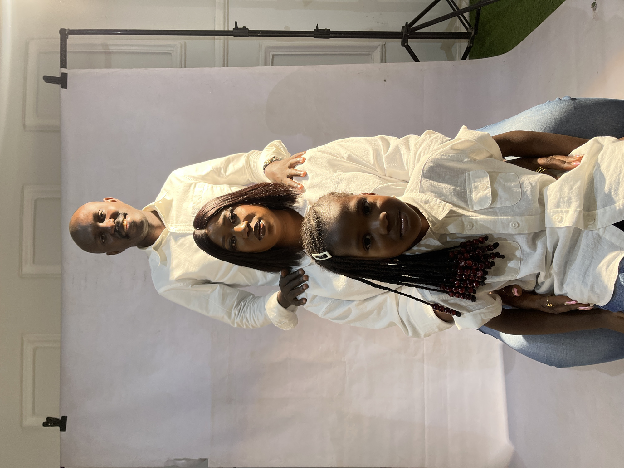 A family portrait features three individuals dressed in white shirts and jeans. An adult male is positioned at the back, followed by an adult female, and a young girl in the front. The background consists of a white backdrop paper roll attached to a stand. An artificial turf is located at the top right corner behind the paper roll. The location is identified as Jos, Nigeria.