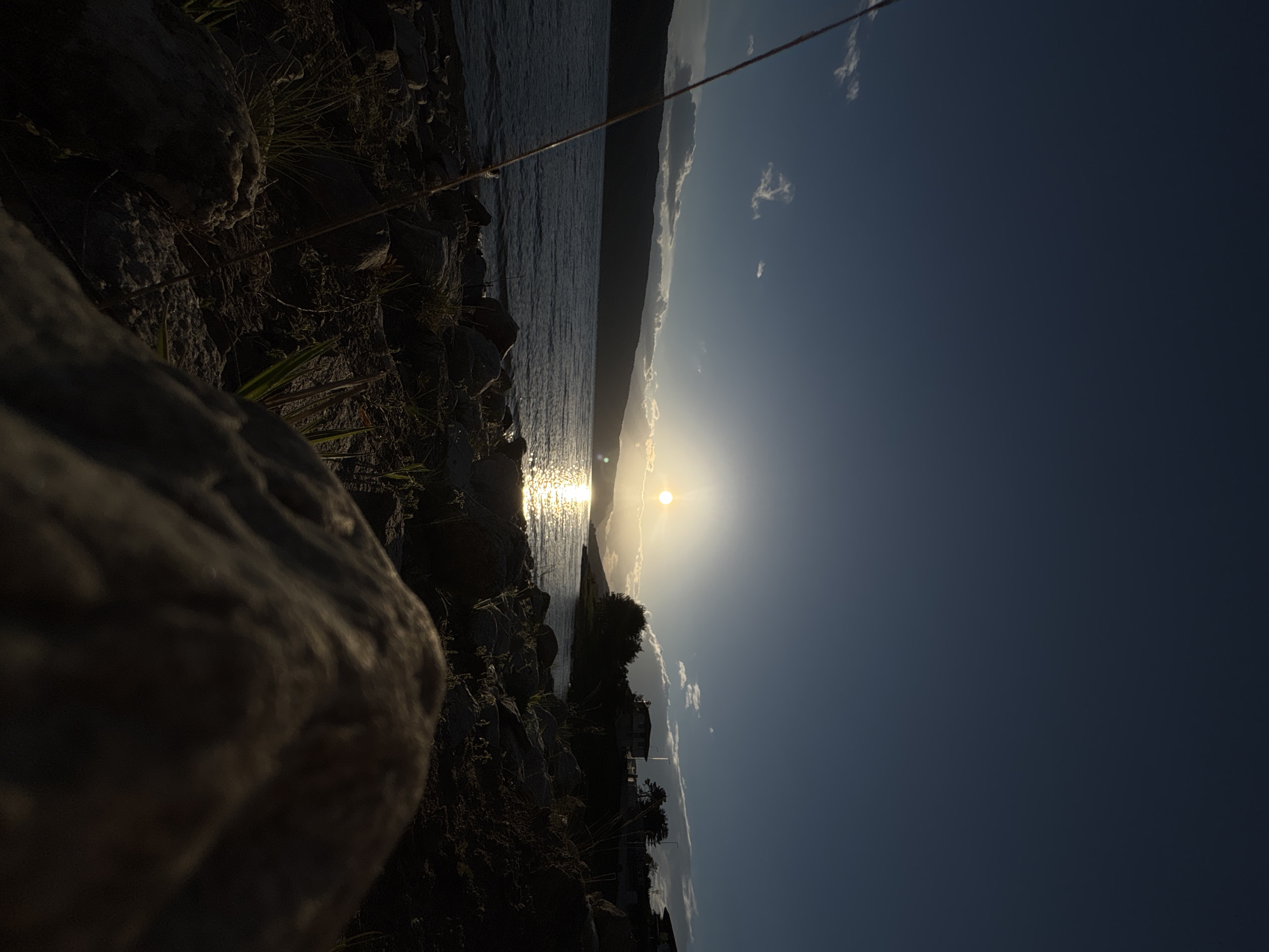 A horizontal shot of a sunset over water, likely a lake or ocean, with a rocky shoreline in the foreground. The sun is low on the horizon, creating a bright reflection on the water's surface. The sky is a gradient of blue, fading from light near the horizon to a deeper shade above. Silhouetted land is visible on the opposite shore. A large rock formation occupies the lower left corner of the frame. Vegetation is visible along the shoreline. There are buildings and trees on the shoreline across the water from the camera.