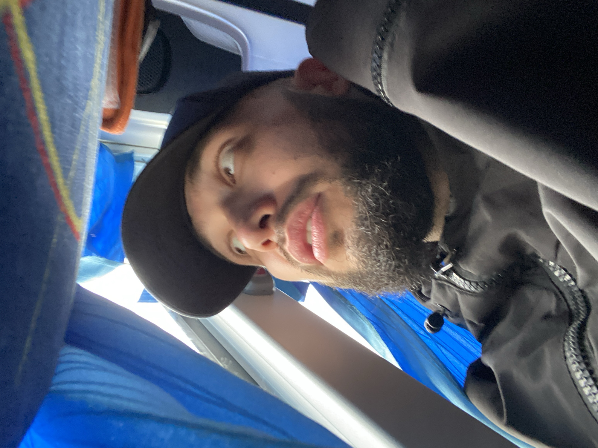 This close-up, low-angle portrait captures a man with a dark beard and mustache, looking upwards and slightly to the side with wide, expressive eyes and a partially open mouth, conveying a playful or surprised emotion. He wears a dark cap or hat and a dark jacket featuring a textured zipper and a small black toggle or cord.
The setting appears to be an interior space, likely a vehicle such as a bus or train, consistent with the provided context of Bereznyaki, Russia. Bright light streams from above and behind him, illuminating various elements of the environment. To the left, there is blue fabric, possibly part of a seat or curtain, with faint vertical yellow and red stripes. More blue fabric is visible in the lower and right sections of the frame. In the upper left background, an orange-colored fabric or object is partially obscured. White structural elements, possibly part of an overhead compartment or window frame, are also visible. The composition suggests a candid moment, possibly a selfie, captured in transit. No visible text is present in the image.