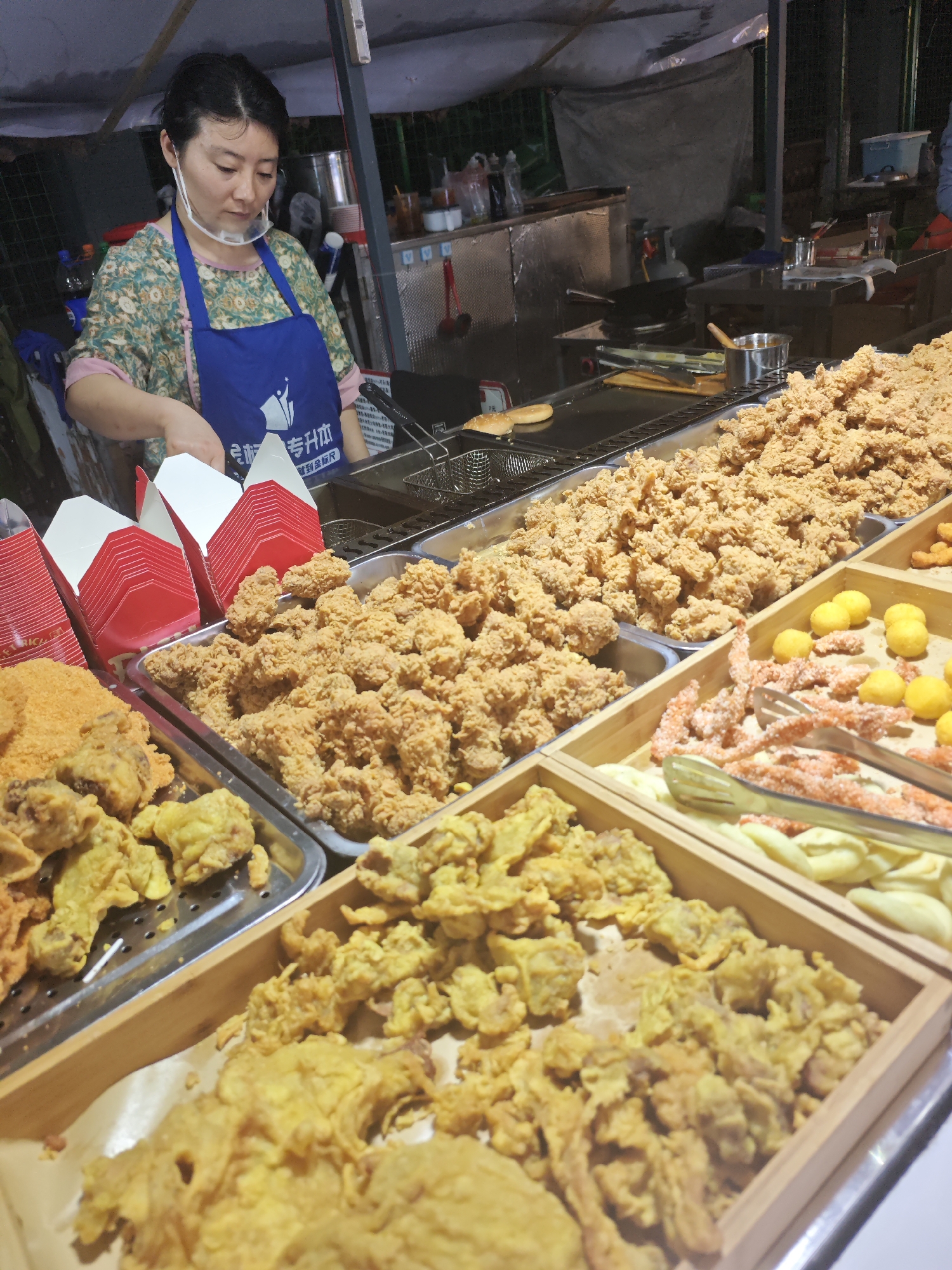 The image depicts a food stall in Shapingba District, China. A woman wearing a blue apron and a face shield stands behind a display of various fried foods, including chicken, other meats, and what appear to be fried balls and shrimp-like items. Red and white takeout boxes are stacked nearby. The woman is attending to food frying in a deep fryer behind the display. The environment appears to be a nighttime food market setting.