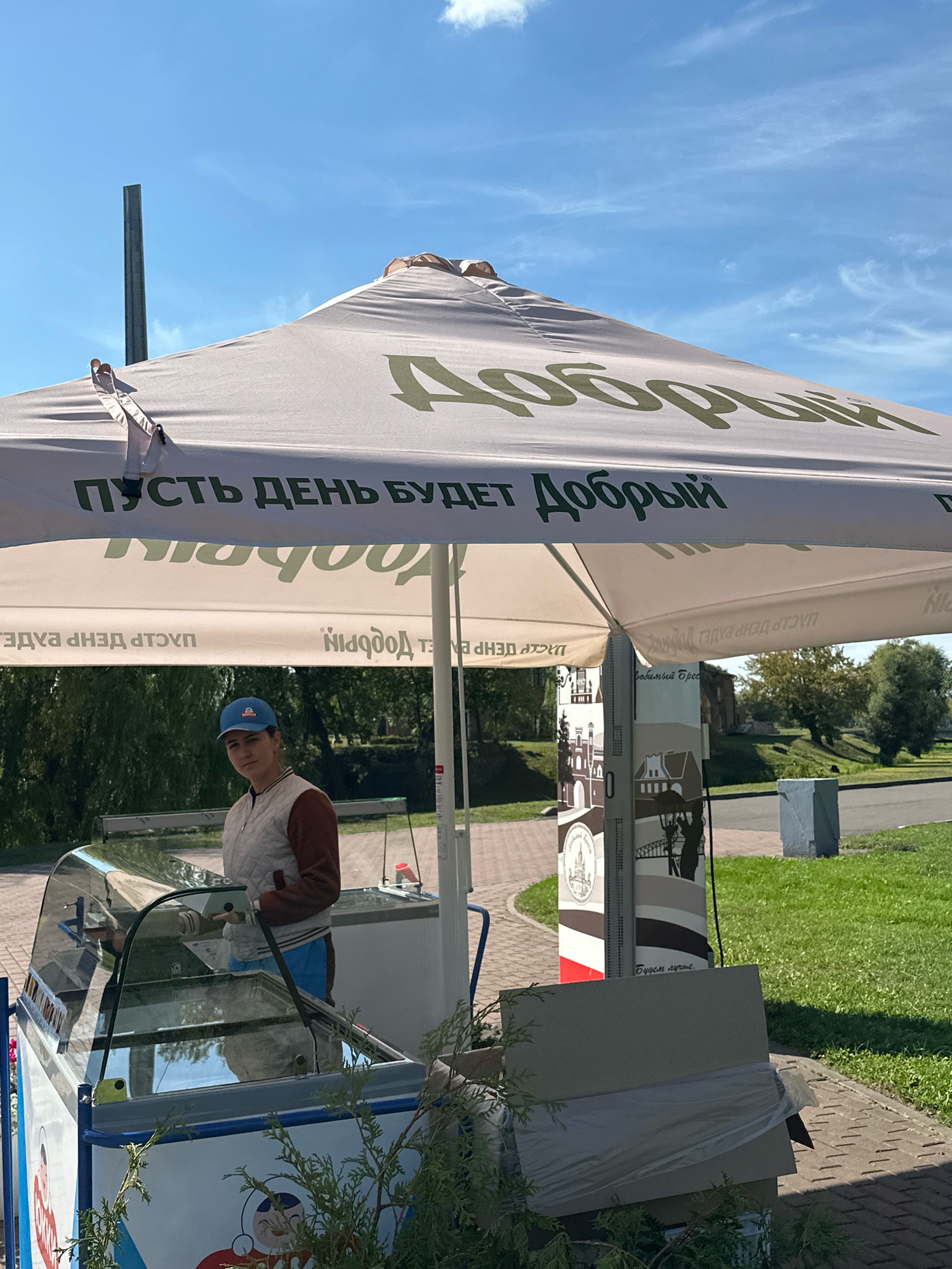 The image shows an outdoor scene under a large umbrella that displays the Russian text "Пусть день будет Добрый" (Let the day be Good) and "Добрый" (Good). A person wearing a blue cap, a beige quilted vest, and a brown shirt is standing behind an ice cream display case. The ice cream display case is illuminated and appears to be open. The background shows a park-like setting with green grass, trees, and a paved walkway. In the distance, a building with an arched window can be seen, possibly part of an old structure or church. The sky is blue with scattered clouds. The overall impression is that of a sunny day with a vendor operating an ice cream stand. The text on the umbrella and the style of the ice cream stand suggest it might be part of a branded ice cream vendor. Without specific landmarks, pinpointing the exact location within Brest, Belarus, is not possible from this image alone, but the scene is consistent with a public park or promenade.