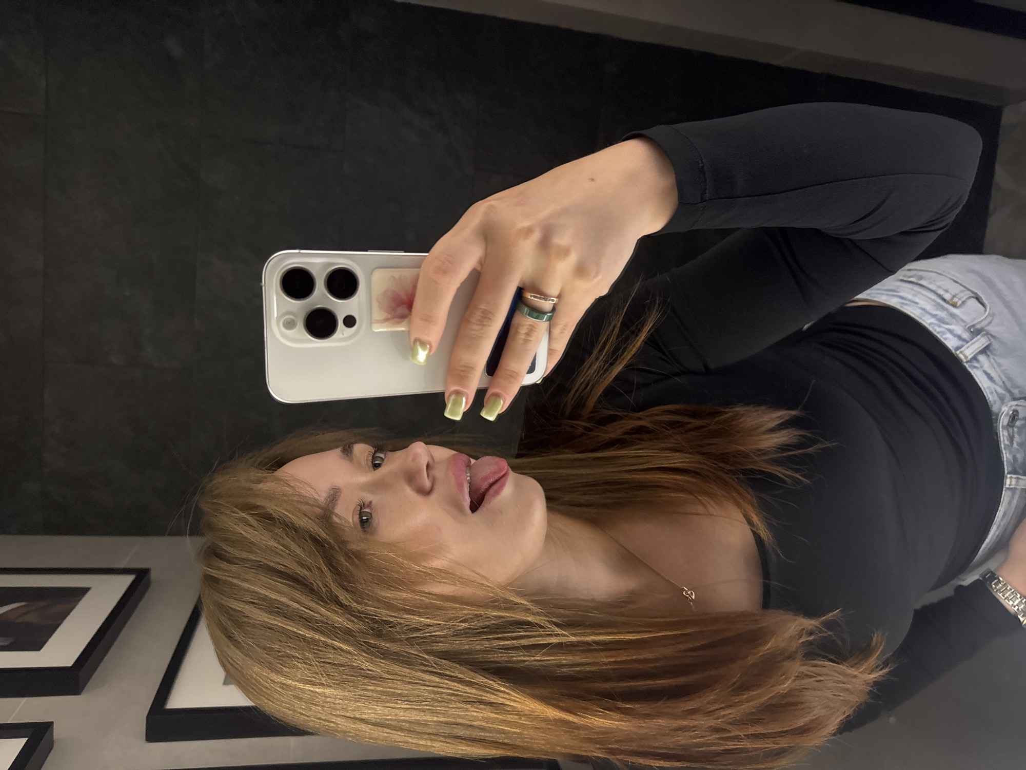In this selfie, a young woman with long, auburn hair is positioned in what appears to be a bathroom. She is looking to her left, with her tongue sticking out playfully. The woman is wearing a black long-sleeved shirt, light-wash jeans, and a silver necklace. She has light green nail polish on her fingernails and is wearing two rings on her right hand. The phone she is using to take the selfie is a white iPhone. The background is a dark, textured wall, with framed pictures visible on the left side of the image. There is a mirror reflection of the woman and the environment. There is no indication of the city of Chaikovsky, Russia in the image itself.