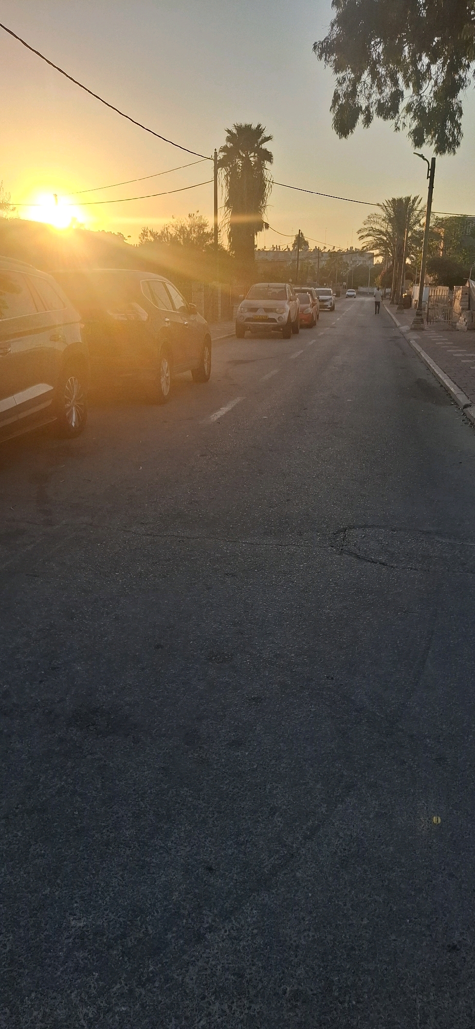 A street scene at sunset in Yerocham, Israel. The sun is low on the horizon, casting a warm glow over the scene and creating lens flare. Several cars are parked on the side of the road, and one person is walking down the street. Palm trees and other vegetation line the street, and buildings are visible in the background. The sky is a clear gradient of orange and blue.