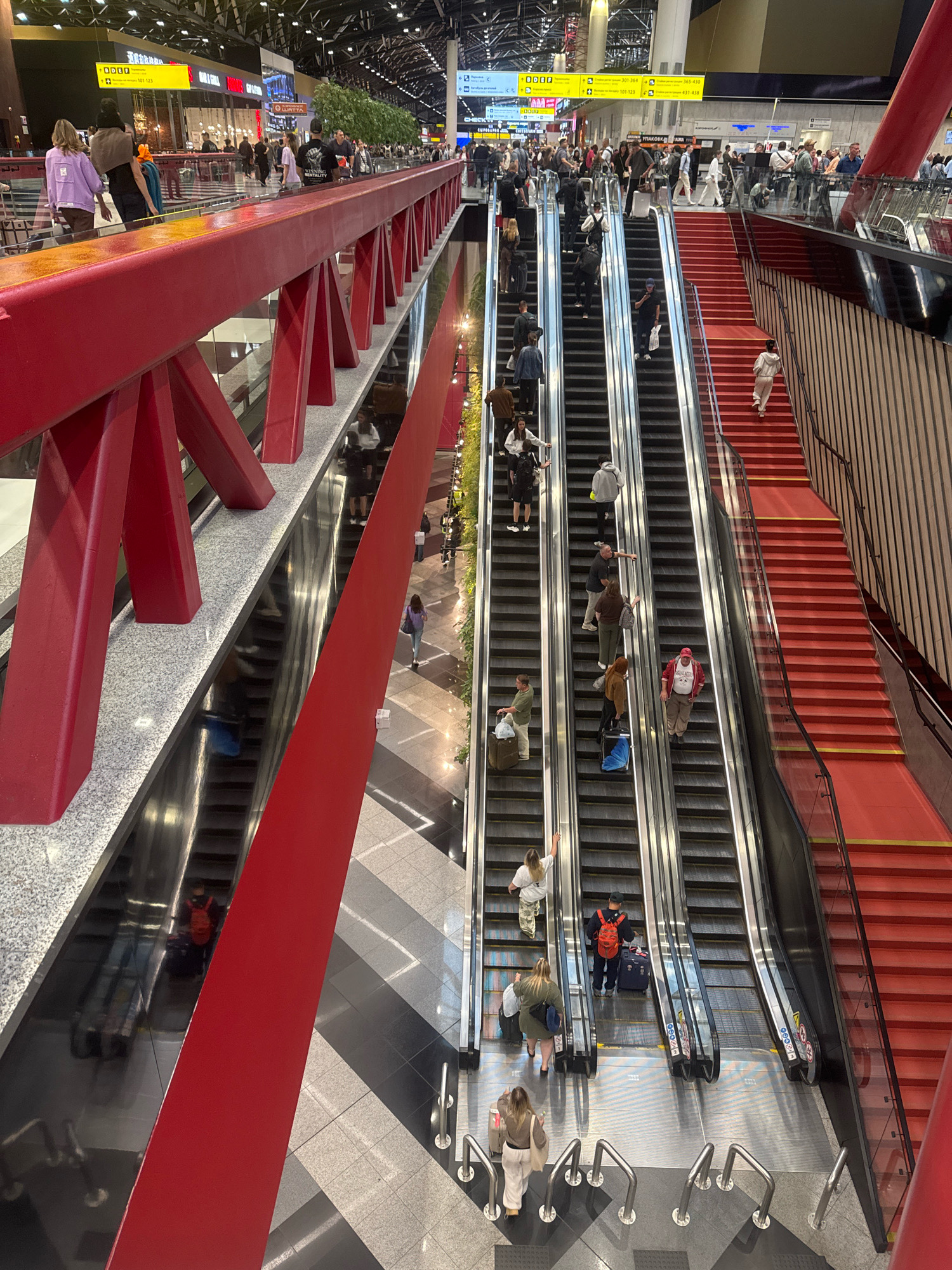 The image depicts a busy interior scene, likely an airport terminal in Khimki, Russia, given the presence of escalators, signage, and a large number of travelers. The architecture features a prominent bright red structure in the foreground, which appears to be a walkway or bridge, with reflective surfaces below. Multiple escalators are visible, some moving upwards and others downwards, carrying passengers with luggage. People are dressed in casual attire, and various suitcases and bags are scattered around. The environment is bright and modern, with high ceilings and prominent lighting fixtures. One escalator on the right has bright red steps, contrasting with the metallic grey of the other escalators. The overall impression is one of movement and transit within a large public space.