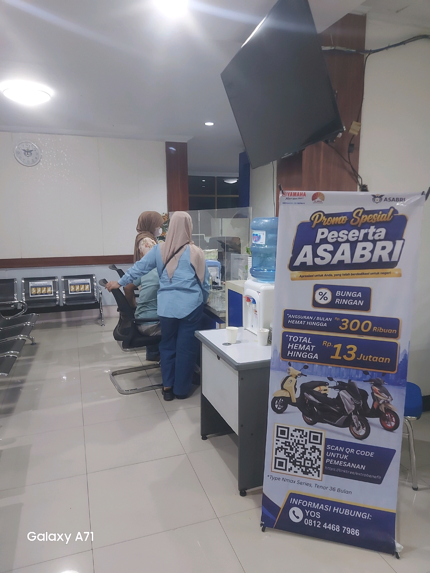 The image shows the interior of a waiting area, likely an office or service center, with a promotional banner on the right side. The banner advertises a "Promo Spesial Peserta ASABRI" (Special Promo for ASABRI Participants), detailing benefits like low monthly installments ("Bunga Ringan" - light interest) of Rp. 300,000 and a total saving of up to 13 million Rupiah. It also features images of motorcycles, a QR code for ordering, and contact information.
In the background, there are rows of dark chairs, a wall clock, and two people, both women wearing hijabs, sitting and standing. One woman, in a light blue long-sleeved shirt and dark pants, is standing and appears to be interacting with someone or something out of frame. The other woman, in a patterned top and a light brown hijab, is seated. There is a water cooler on a stand, and office desks or counters are visible. The flooring is tiled in a light neutral color. The overall lighting suggests an indoor, possibly artificial, illumination. The "Galaxy A71" watermark is present at the bottom left of the image, indicating the device used to capture the photo.