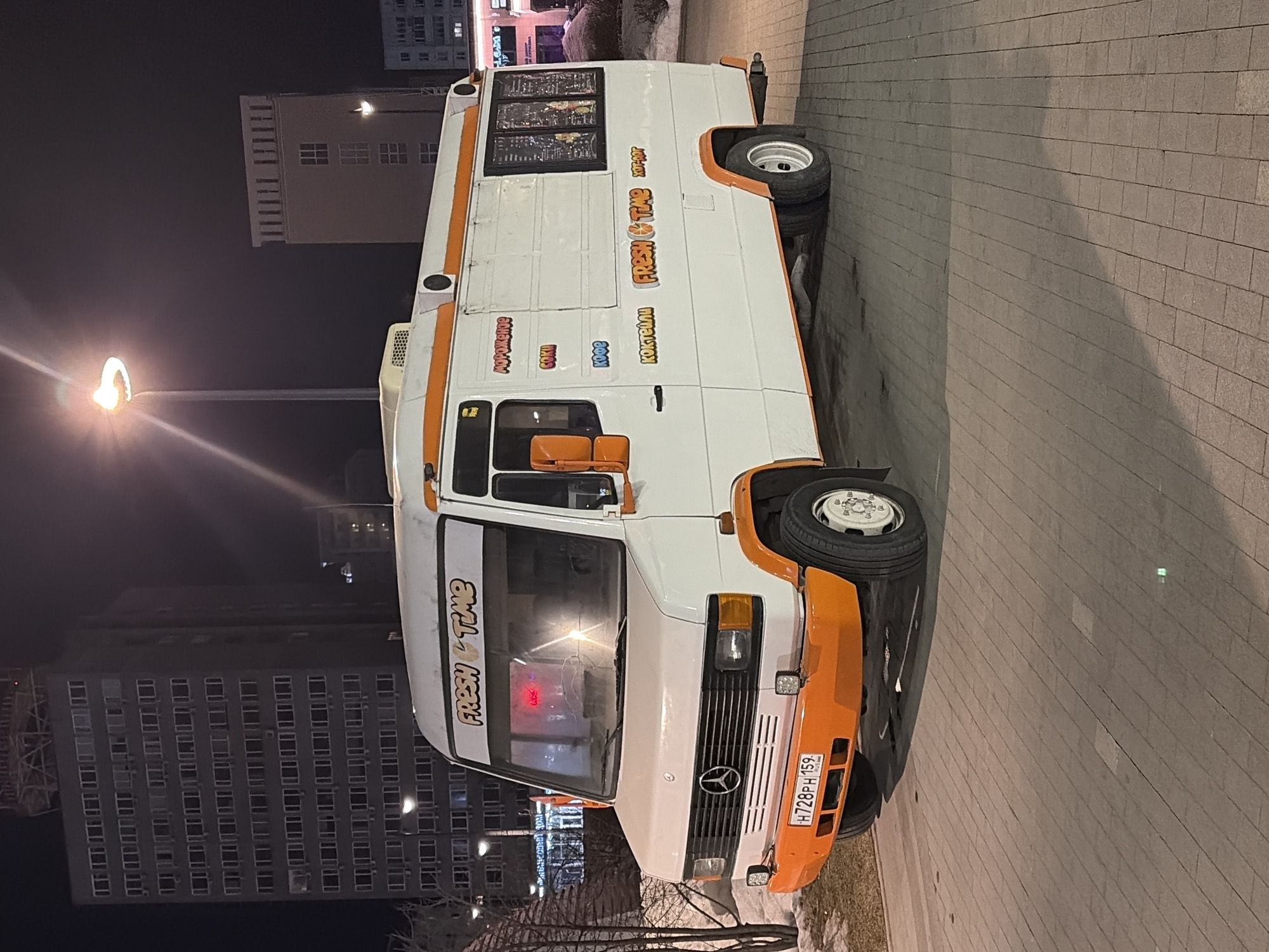 A white and orange Mercedes-Benz food truck, identified by its license plate H728PH 159 (Perm Krai region), is parked on a patterned brick-paved surface during nighttime. The vehicle is branded "FRESH & TIME" and lists menu items in Russian on its side, including "МОРОЖЕНОЕ" (Ice Cream), "СОКИ" (Juices), "КОФЕ" (Coffee), "КОКТЕЙЛИ" (Cocktails), and "ХОТ-ДОГ" (Hot Dog). A bright streetlamp to the right illuminates the scene, casting a lens flare. In the background, a tall, dark grey multi-story building, identified as the Hotel Ural at ulitsa Lenina, 58 in Perm, Russia, with numerous rectangular windows and a visible rooftop structure, stands prominently to the left. Further to the right, a lighter beige building with vertical facade elements is partially visible. Patches of snow and bare tree branches are present on the left foreground. No individuals are visible in the scene.
