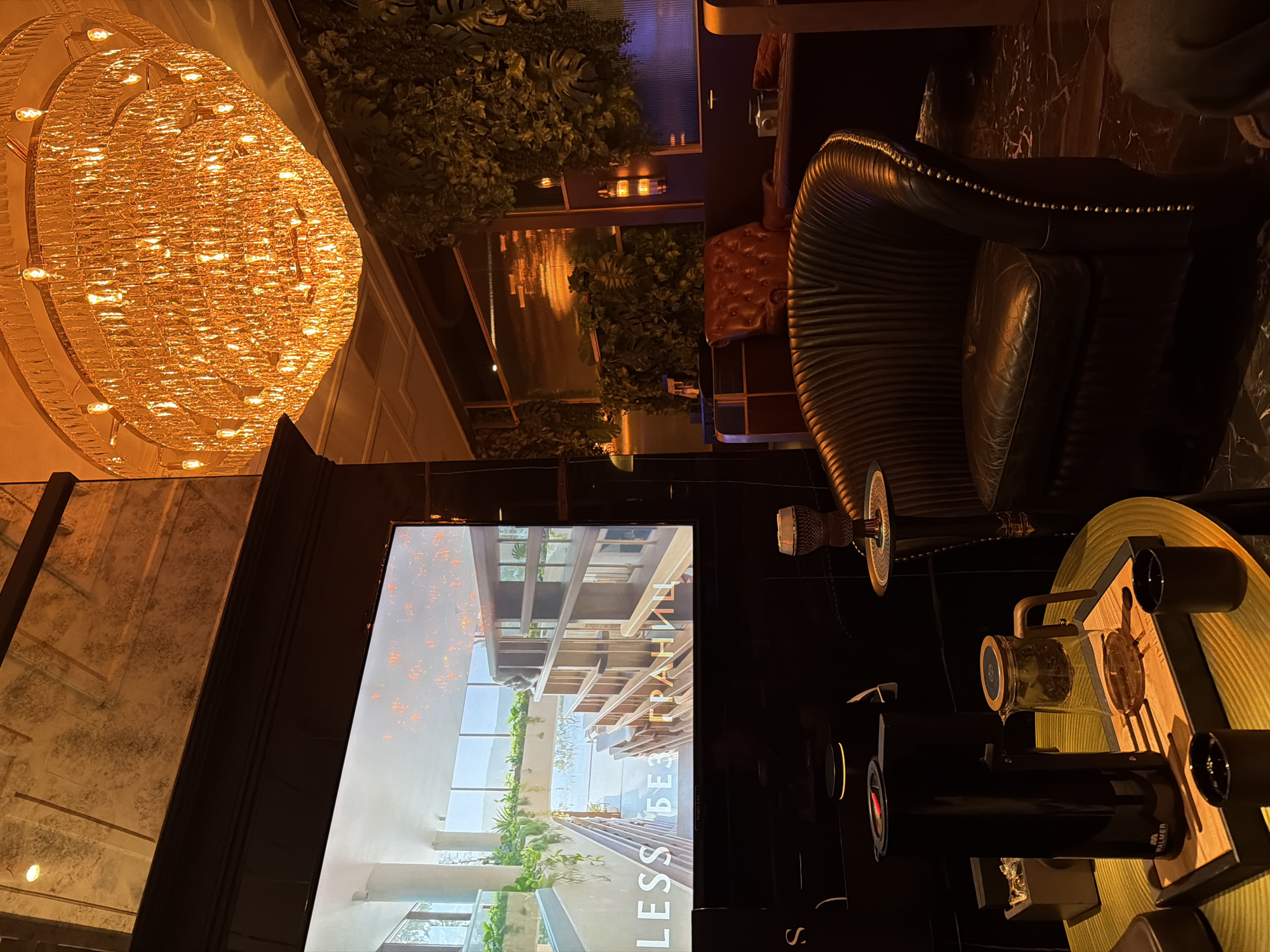 A low-angle indoor shot shows a brightly lit interior, perhaps a luxury bar or lounge in Moscow, Russia. A large, ornate chandelier dominates the upper left, casting a warm, orange glow. The background features greenery, suggesting a rooftop or open-air space.
In the foreground, a dark leather chair is visible, indicating a seating area. A table displays a complex arrangement of objects, possibly a hookah or smoking apparatus. A large television screen shows an architectural image, with the Cyrillic text "БЕЗ ГРАНИЦ" (without borders) visible.