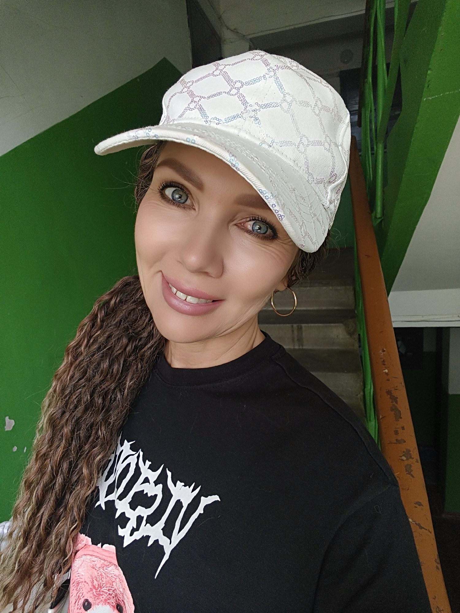 In a medium shot, a woman with blue eyes and brown curly hair tied in a ponytail is smiling. She is wearing a white baseball cap with a patterned design and a black t-shirt with a white graphic. The background is a green painted wall and a wooden railing next to a staircase. The lighting suggests an indoor, possibly residential, setting.