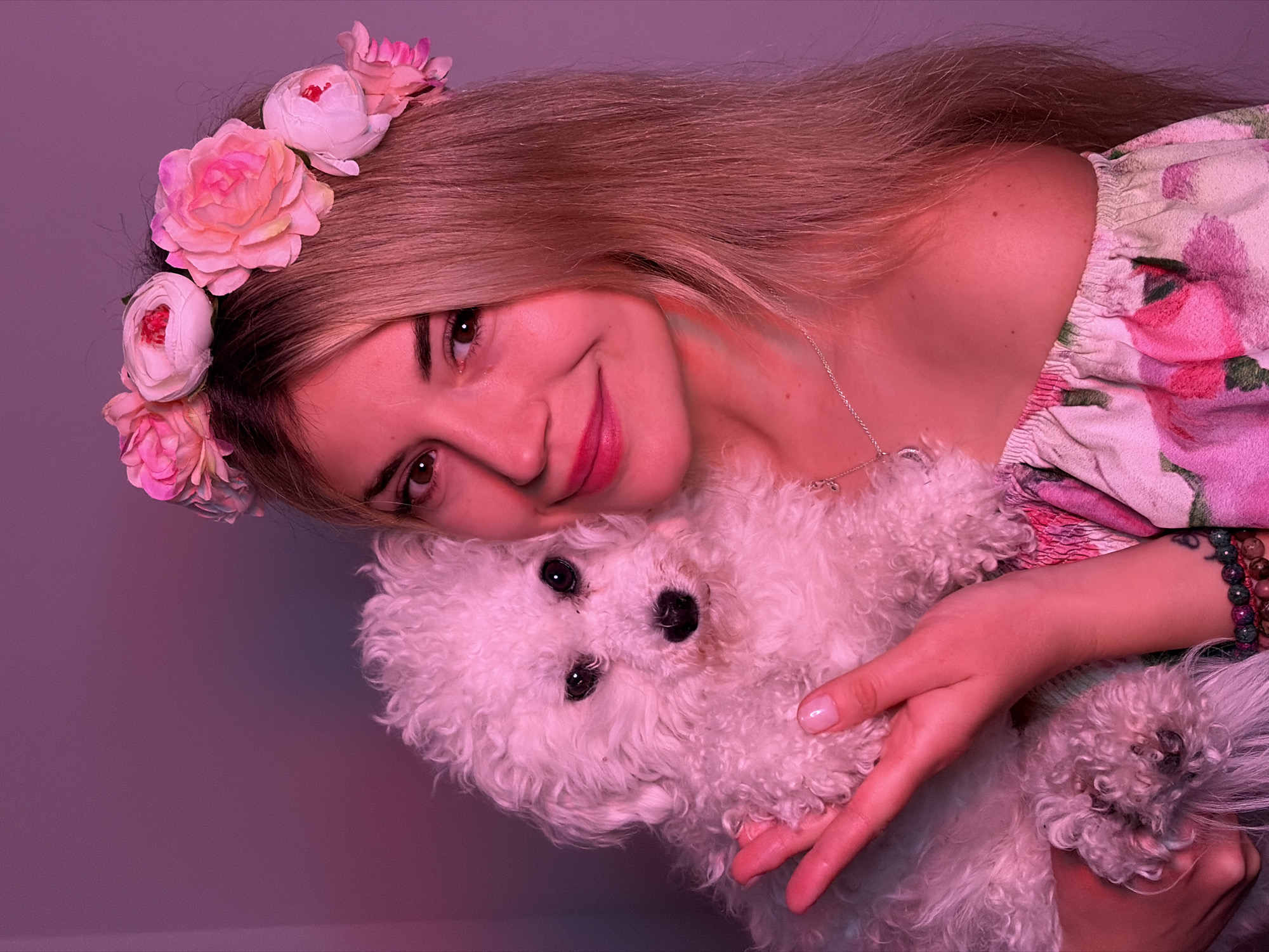 A young woman with long blonde hair wearing a floral headband and a floral-patterned top is hugging a small white dog. The woman is smiling. The background is a gradient of light purple to darker purple. A bracelet is visible on her right wrist, and a thin necklace is visible around her neck. The scene is under a strong pinkish-purple light. The location is not possible to determine from the image.