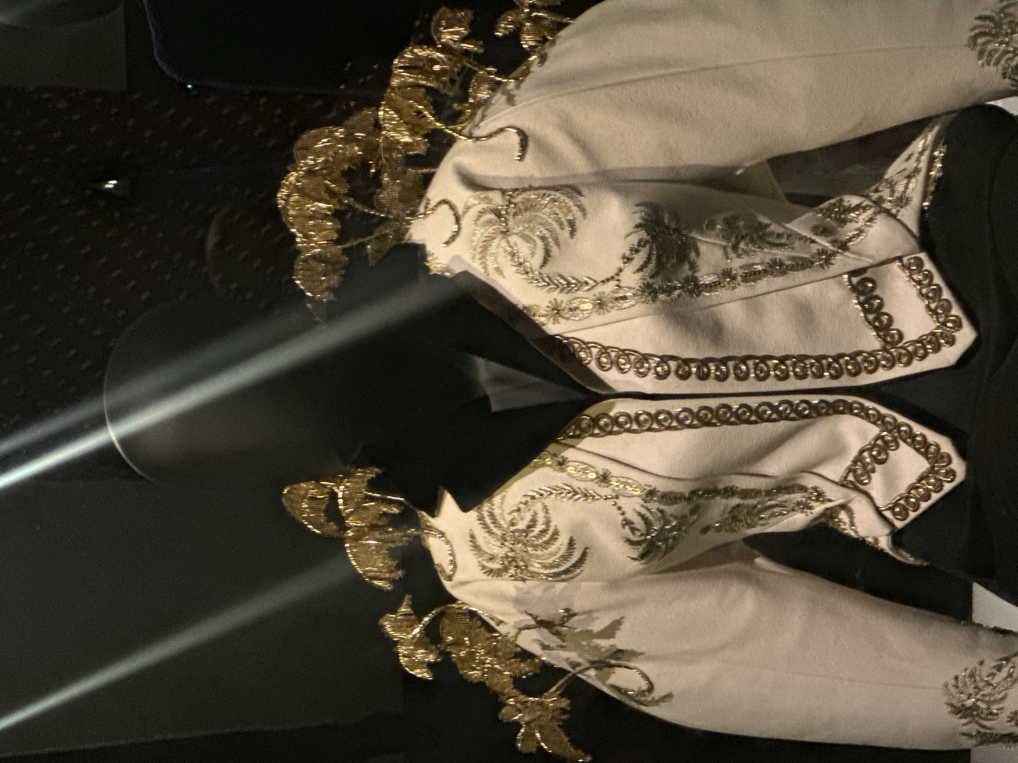 The image displays a cream-colored, heavily embroidered jacket on a black, headless mannequin. The jacket features black pointed lapels and is adorned with intricate gold thread embroidery, primarily depicting stylized palm fronds or botanical patterns across the front panels and sleeves. Vertical lines of gold circular embellishments run along the front opening and bottom hem of the jacket. Elaborate, three-dimensional golden structures, resembling clusters of leaves or small trees, are attached to the shoulder areas, extending upwards. The mannequin is positioned against a dark background within a display environment, likely a museum or exhibition in London, United Kingdom, characterized by bright, directional overhead lighting casting prominent reflections across the upper portion of the frame.