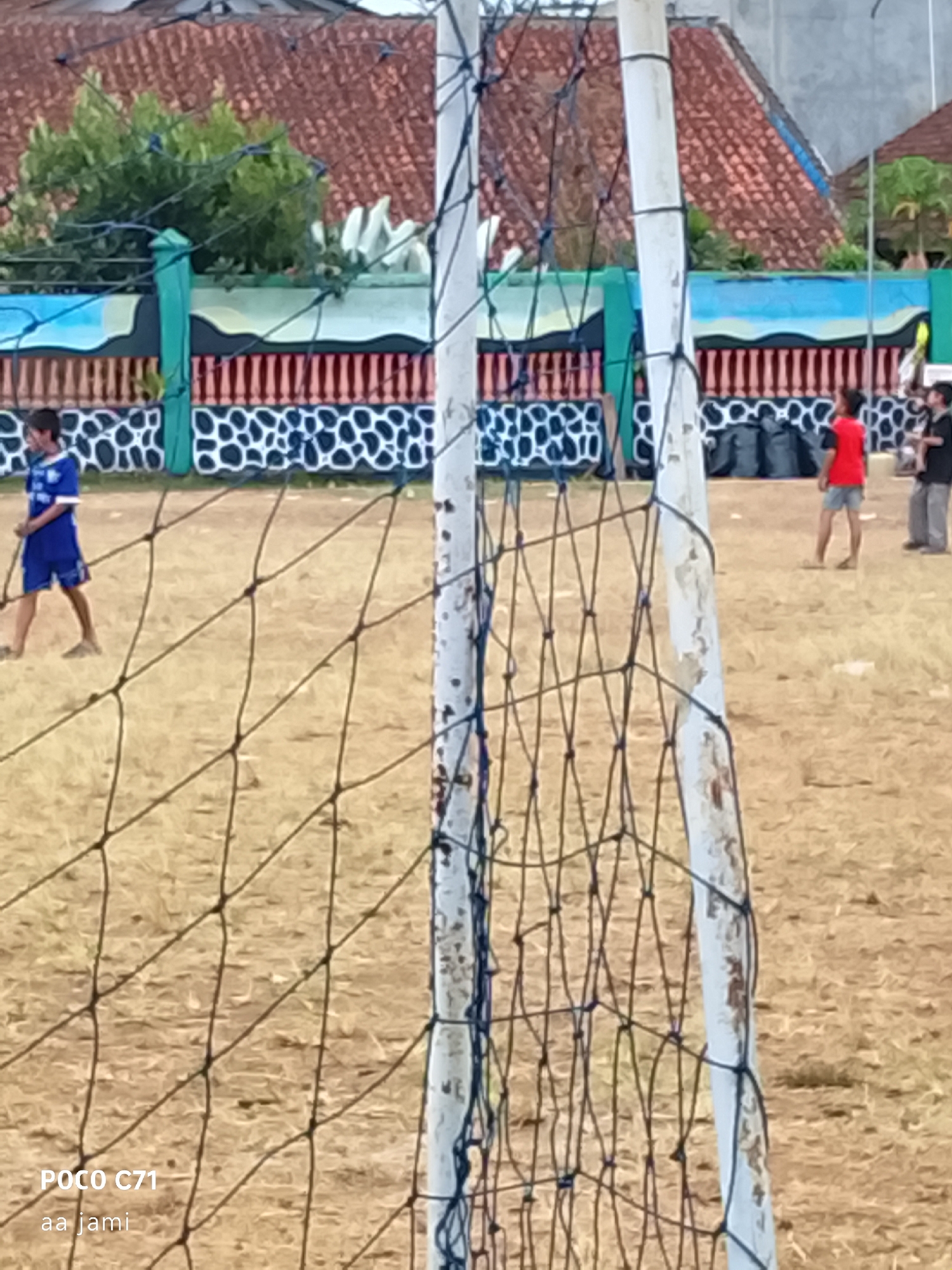 In Tasikmalaya, Indonesia, a group of children are playing soccer on a dry, dirt field. In the foreground, a soccer net is visible, partially obscuring the view. A young boy in a blue soccer uniform is dribbling a ball to the left. Further back, a boy in a red shirt is standing, and a man in a black shirt and grey pants is also present. A colorful mural adorns a wall in the background, with a tiled roof visible above it. The overall scene depicts an informal soccer game in a somewhat dusty environment.