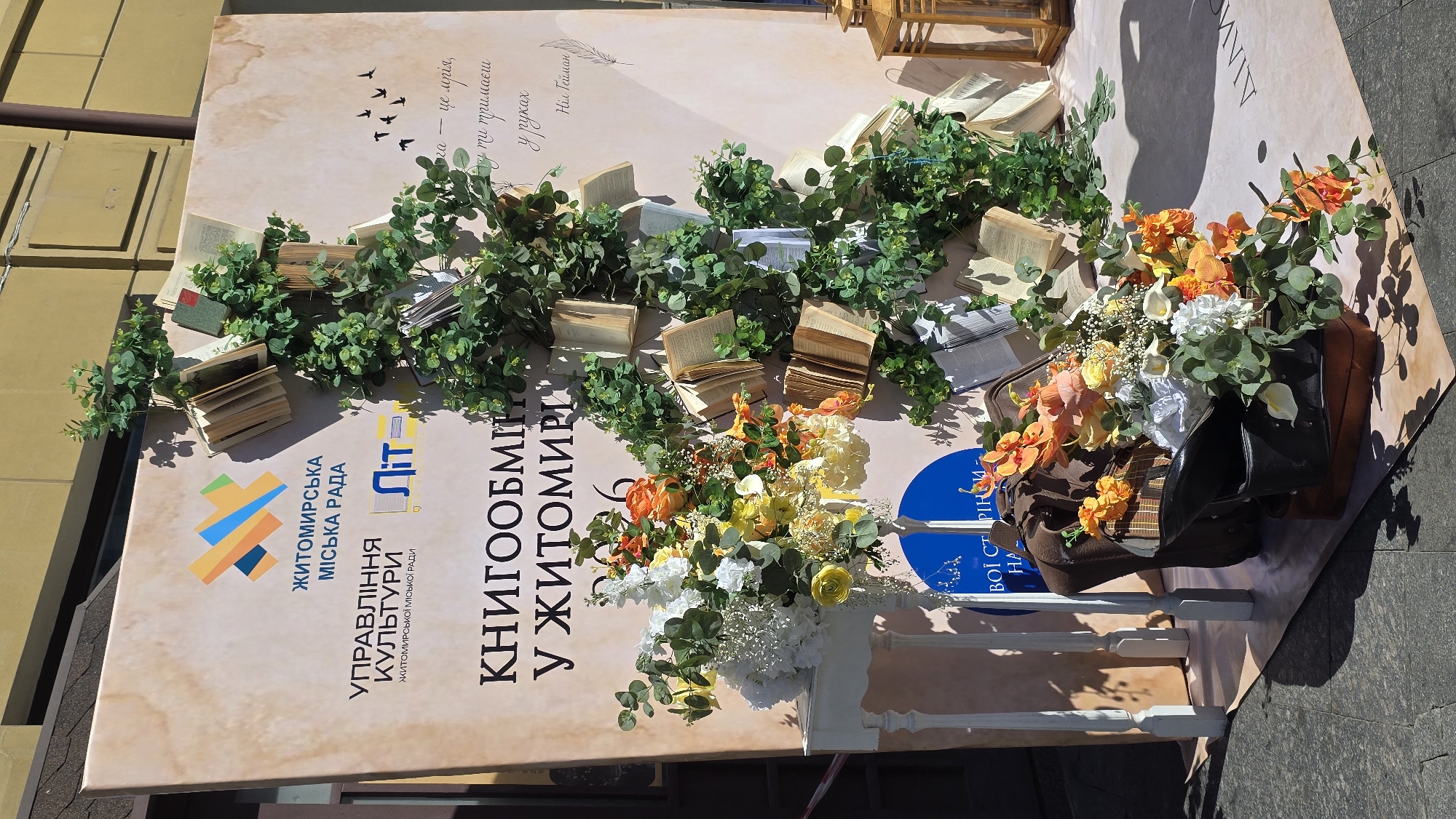 The image shows an outdoor decorative display in Zhytomyr, Ukraine, likely for a cultural event. The focal point is a large banner with text in Ukrainian, featuring logos and graphics. A vertical arrangement of greenery, adorned with open books, cascades down from the top right of the banner. To the left of the banner, a white decorative stand holds a large floral arrangement in shades of orange, yellow, and white, with eucalyptus leaves. In front of the stand, there are two vintage suitcases, one open and overflowing with flowers. The display is set against a building facade, with shadows cast on the ground, indicating it is daytime and sunny. The overall aesthetic is whimsical and literary, celebrating books and nature.