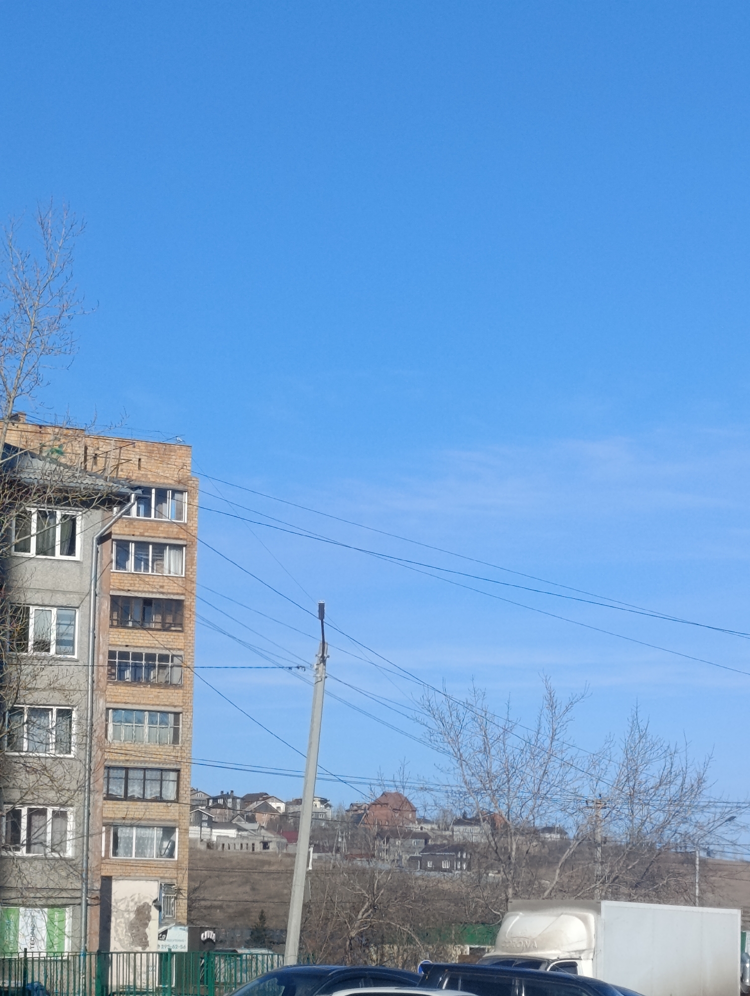 The image depicts a building facade in Krasnoyarsk, Russia. The building, located on a street with utility poles and power lines, features a combination of gray and brick sections. A fence is visible in the foreground. In the background, other buildings appear on a raised landform or hill. Several cars and a truck are parked nearby. The sky is clear and blue.