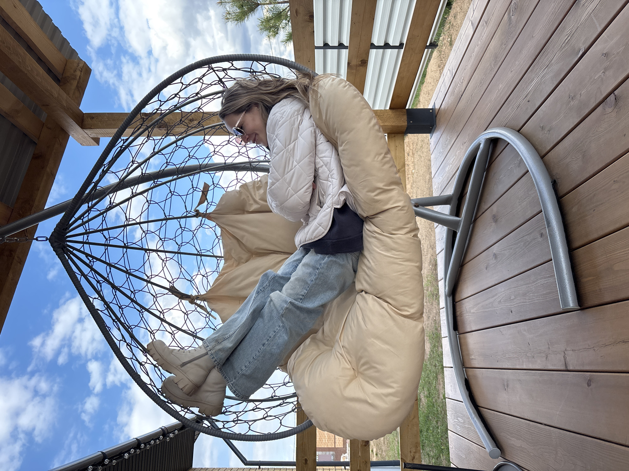 A young person wearing a white puffer jacket, blue jeans, and beige boots is sitting in a net chair with a beige cushion in what appears to be an outdoor setting. The chair is suspended from a metal frame. The sky is visible through the net of the chair, suggesting an outdoor location. The setting includes wooden structures and a roof in the background. This photo was taken in Chaikovsky, Russia.