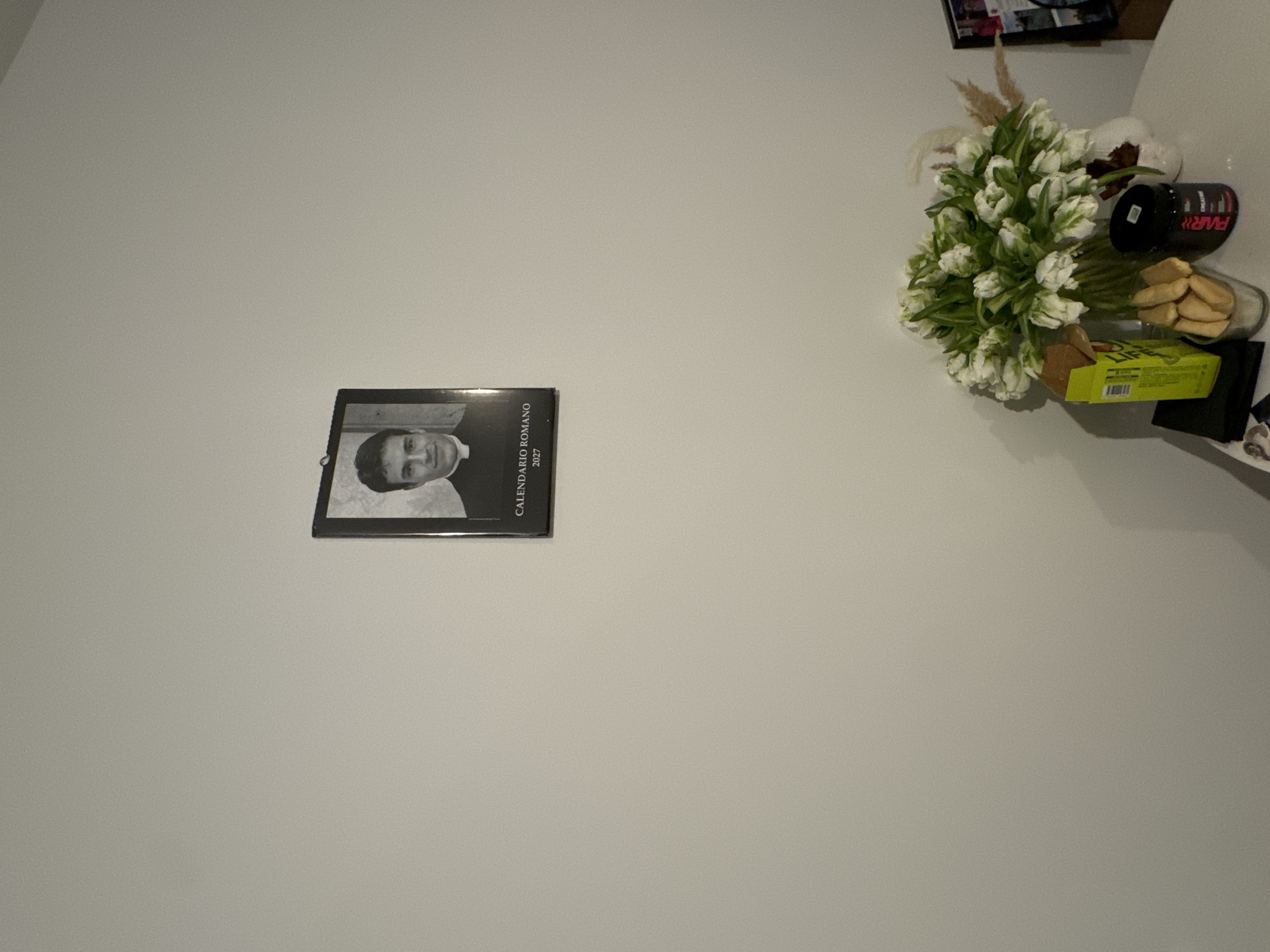 The image shows a plain white wall with a calendar and a collection of items on a surface.
On the wall, to the left and slightly below the center of the frame, hangs a black and white calendar. The calendar features a portrait of a man in clerical attire. The text "CALENDARIO ROMANO 2027" is visible on the calendar.
On the right side of the frame, a white surface holds a cluster of objects. These objects include:
- A bouquet of white tulips.
- A white decorative object.
- A black container of what appears to be a nutritional supplement.
- A glass jar containing cookies.
- A yellow and green box.
- A black object of indeterminate shape.
The image provides no information related to a location in Moscow, Russia.