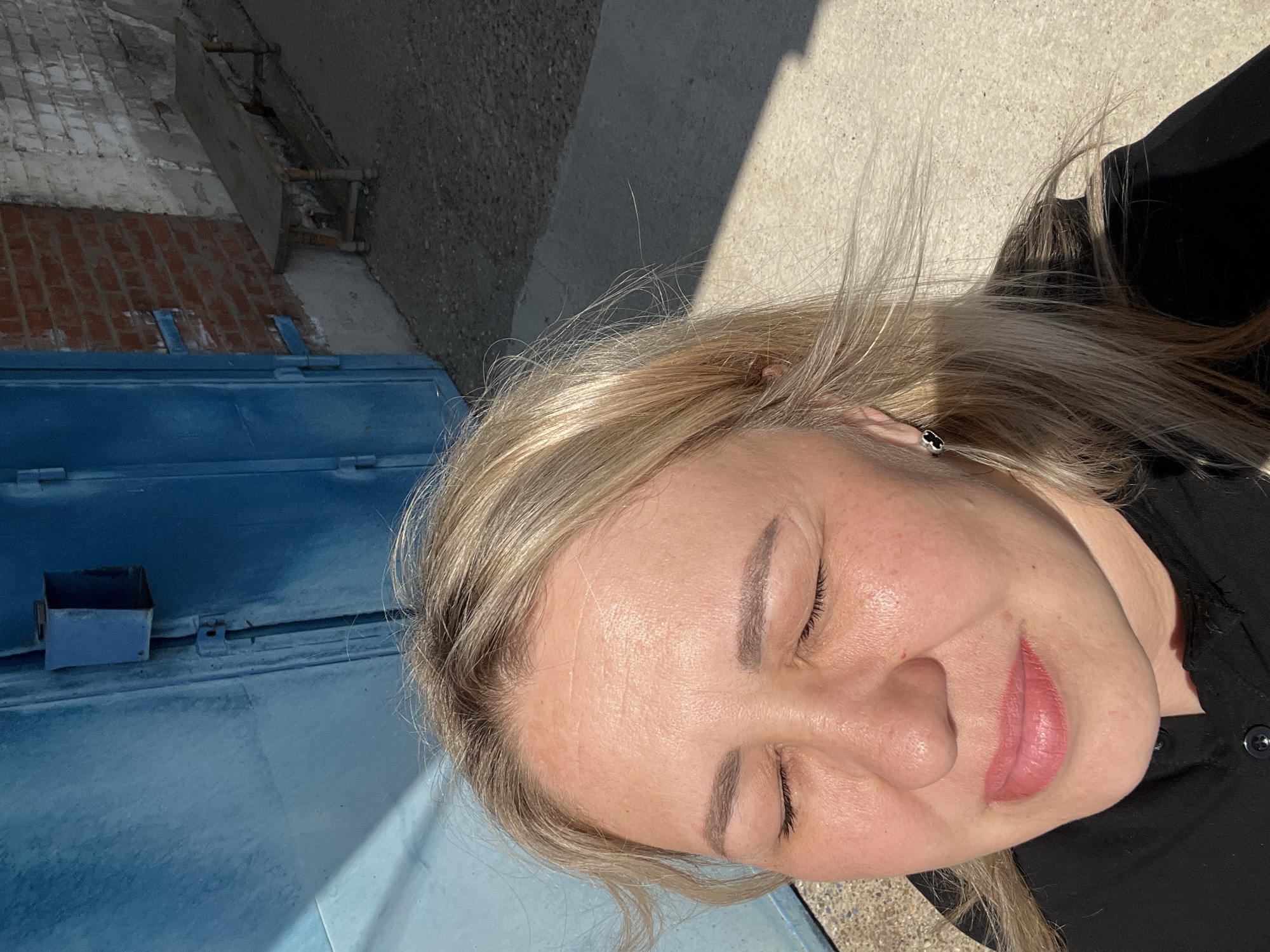 A close-up, slightly rotated, outdoor selfie of a blonde woman with her eyes closed. She has fair skin, a small mole on her forehead, and pink lipstick. She's wearing a black top. Part of a blue metal object is visible to the left. Behind the woman is a grey concrete surface and a brick building fragment. The location is unknown; there is no definitive indication that the image was taken in Chaikovsky, Russia.
