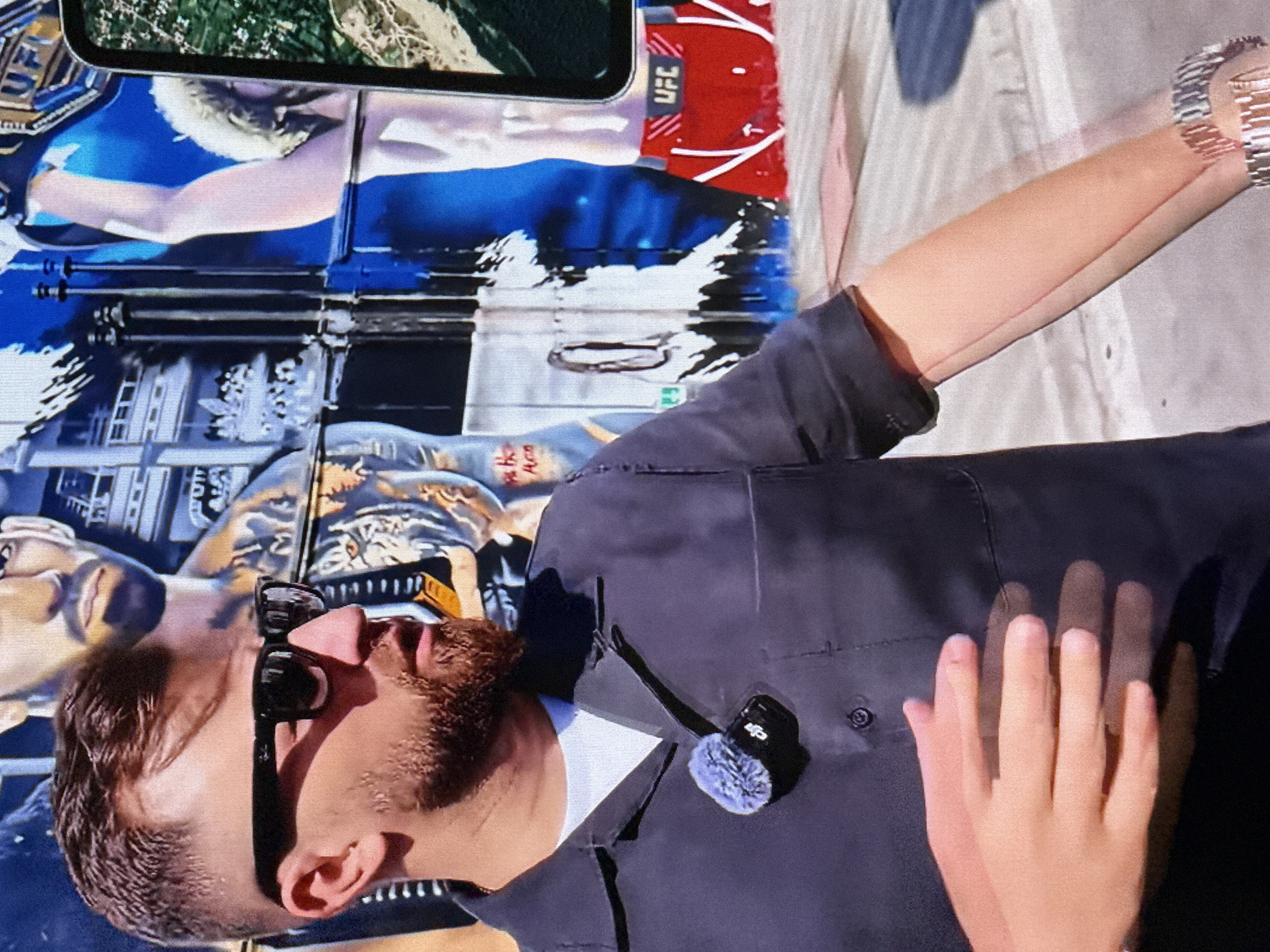 A man wearing a dark blue collared shirt, a white t-shirt, and black sunglasses is standing in front of a mural. He has a reddish-brown beard and short brown hair. A small furry microphone is clipped to his shirt. His hands are in front of him, with his left hand resting on his right wrist. On his right wrist, he is wearing a silver watch. The mural behind him depicts two figures. The figure on the left is a man with a beard and tattoos. The figure on the right is a woman with blonde hair wearing red UFC shorts. The mural is colorful, with blues, whites, and reds. The ground in front of the mural appears to be concrete.