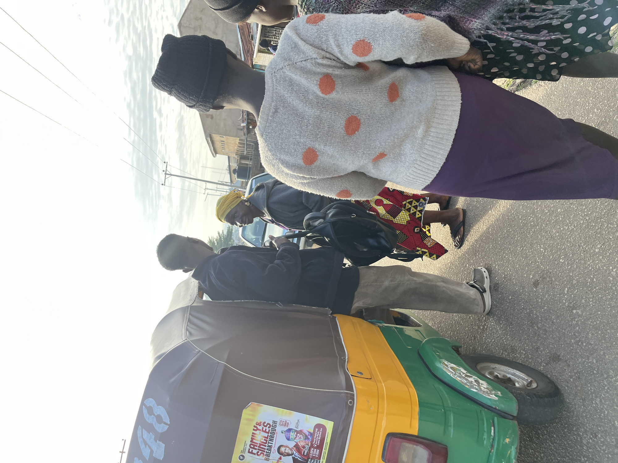 In the city of Jos, Nigeria, on a paved road with some debris, a group of people stand near a yellow and green auto-rickshaw. A young man in a dark jacket and light-colored pants talks to a woman wearing a yellow headscarf and a colorful patterned skirt. Another young person, facing away, wears a white sweater with orange polka dots and a dark beanie. Behind them, another individual is partially visible. The auto-rickshaw has a brown canopy and a poster on its side advertising a "FAMILY & SINGLES BREAKTHROUGH" event. Power lines and buildings are visible in the background. The sky is bright and appears overcast.