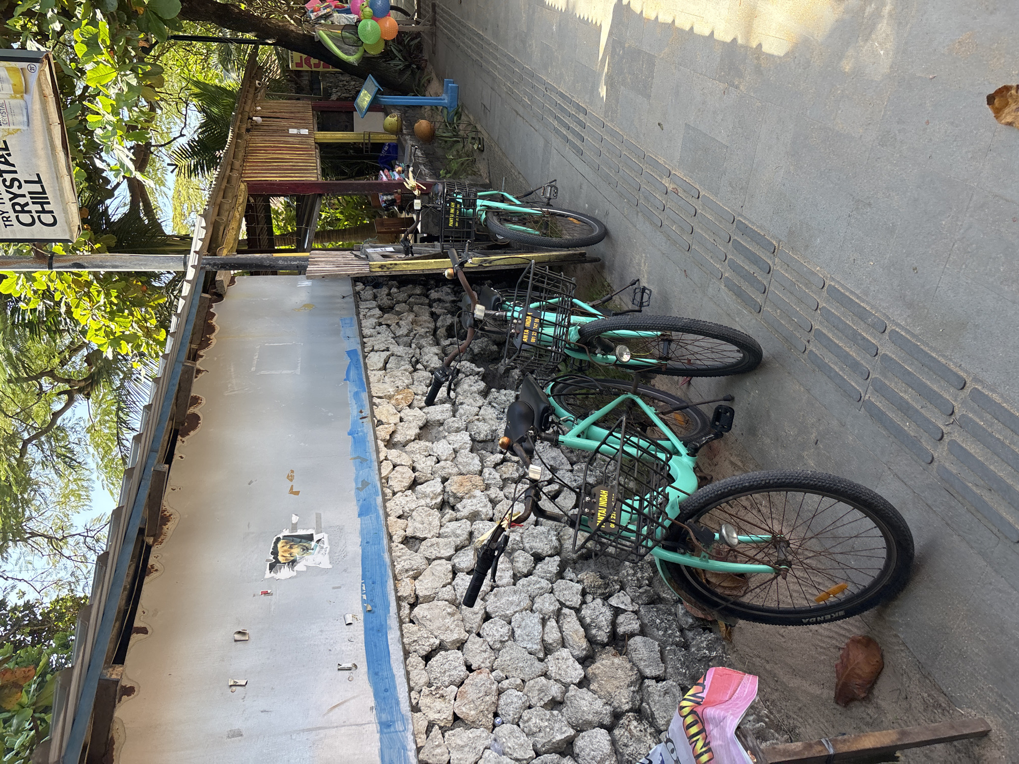 The image shows a street scene in Denpasar, Indonesia. Three light-green bicycles with black baskets are parked against a low stone wall, bordered by a faded building on one side and a tiled sidewalk on the other. The building is a faded white with a blue stripe and remnants of torn posters. The stone wall is made of rough, light-gray rocks. The sidewalk is made of gray tiles with tactile paving. In the background, there's a small wooden structure, possibly a shop or stall, with various items on display, including balloons and coconuts. A sign that reads "TRY CRYSTAL CHILL" is visible. The scene is outdoors during daylight, with shadows cast on the sidewalk. The overall impression is of a relaxed, possibly tourist-oriented, area.