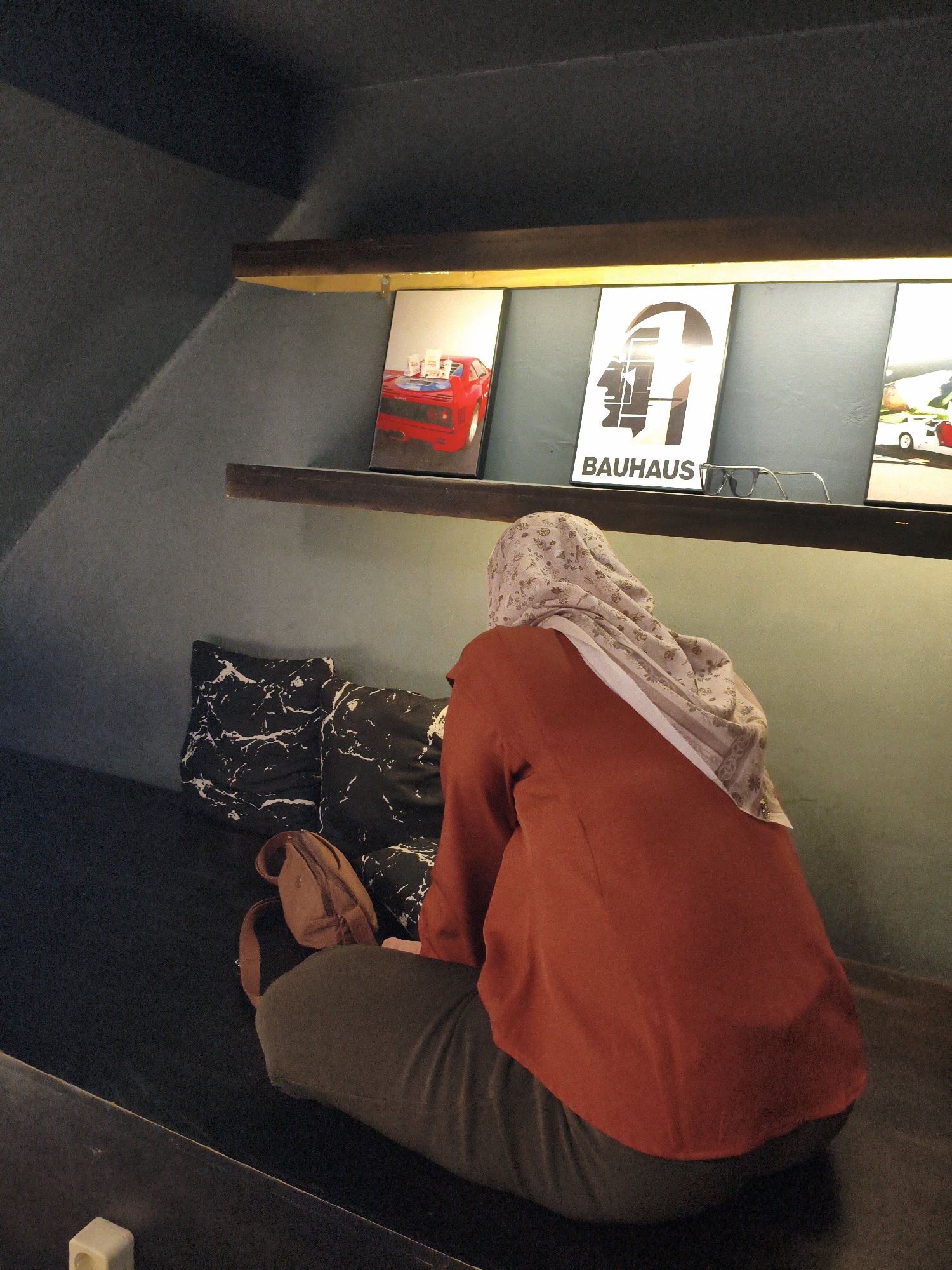 A person wearing a hijab and an orange jacket sits on a dark couch with two black and white patterned pillows. A brown bag rests on the couch next to them. The person is facing away from the camera, looking at a shelf above them. On the shelf are three framed pictures, one of a red car, another with the word "BAUHAUS" and a graphic of a face, and a third of a white car. The walls are a muted green and dark gray, and the lighting is dim. The scene appears to be indoors, possibly in a cafe or a private dwelling. The location is the city of Tebing Tinggi, Indonesia.