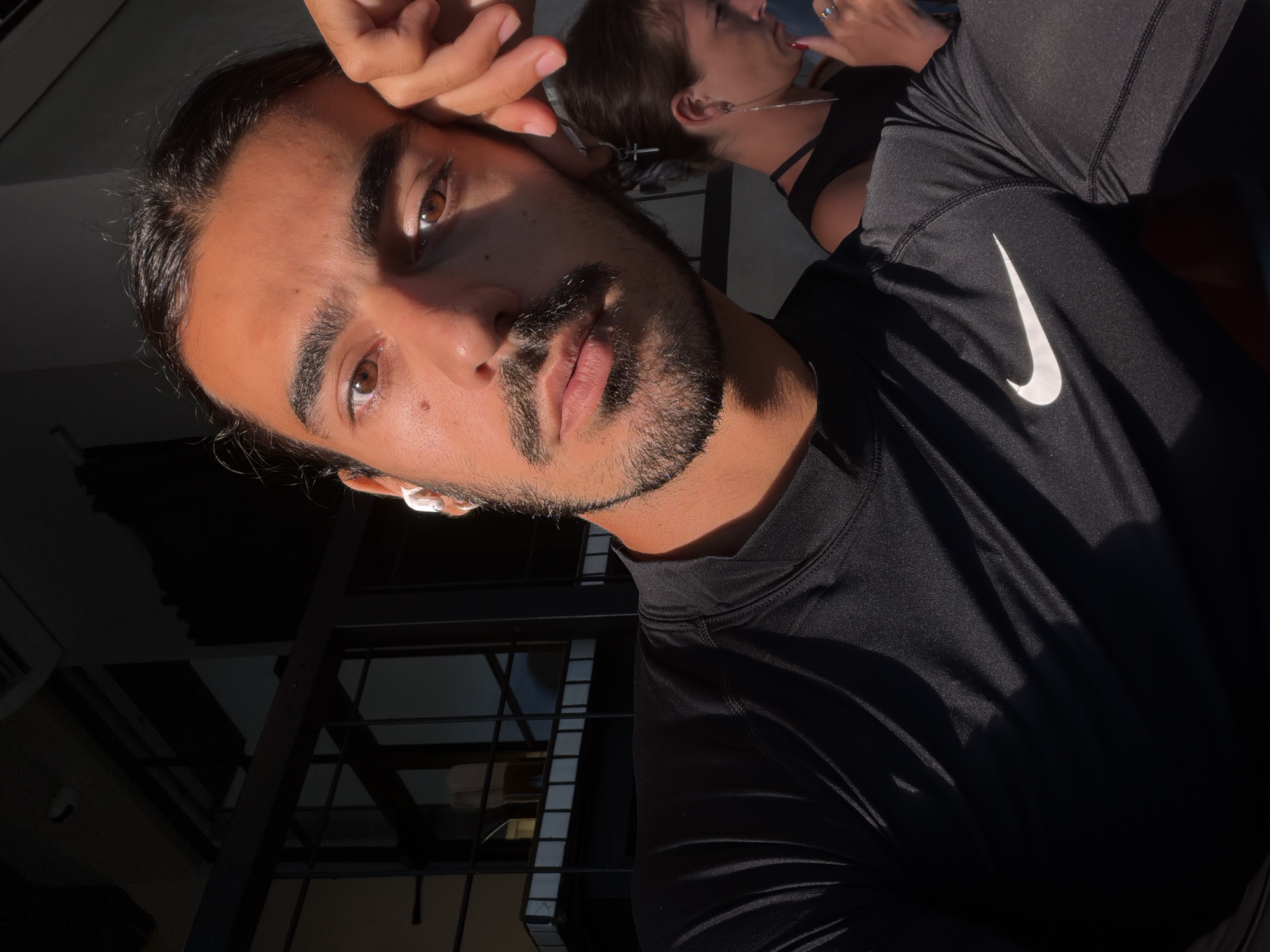 A close-up, angled selfie shows a man with dark hair pulled back, a trimmed beard, and brown eyes. He is wearing a black Nike long-sleeved athletic shirt and an AirPod in his left ear. His right hand is touching his forehead. The background is partially visible, including a woman with her hair up, a black top, and silver necklaces. There is a metal structure similar to a security gate or part of a building. Due to the partial view of the background, a specific location in North Kuta, Indonesia cannot be determined.