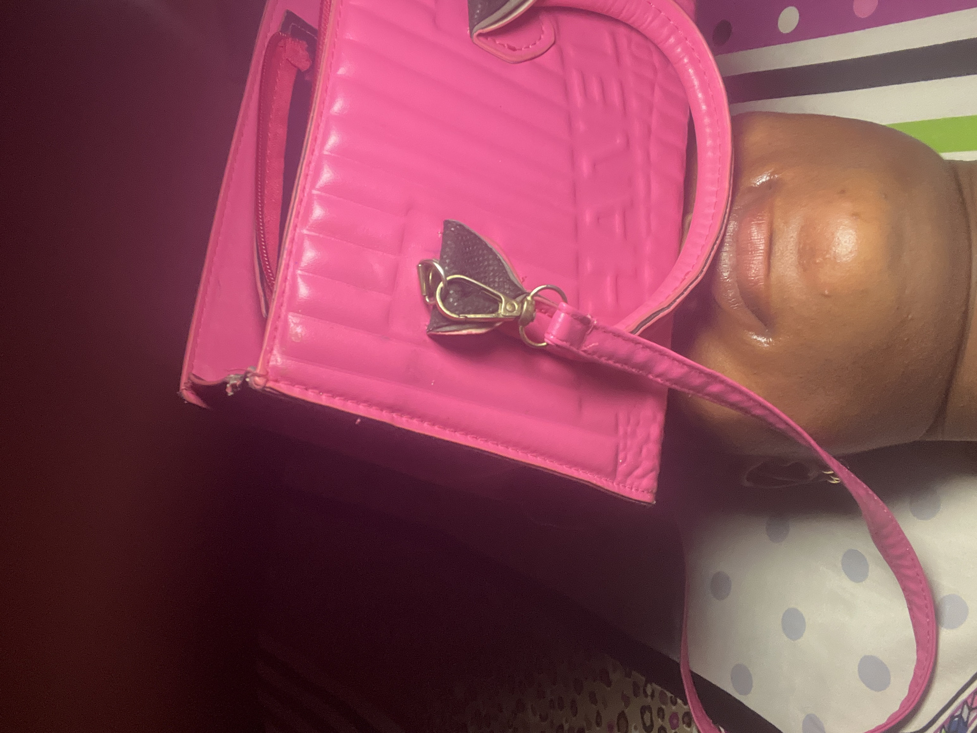 A close-up, angled shot shows a hot pink, faux-leather handbag with a handle and detachable shoulder strap. The bag is partially over a person's face, specifically the cheek and lips. The person is lying on a surface with a white background and light blue polka dots, interspersed with striped sections in purple, white, black, and green. The lower left corner of the image is dominated by dark, blurry shadows. There's insufficient visual information to determine the precise location within Ado, Nigeria.