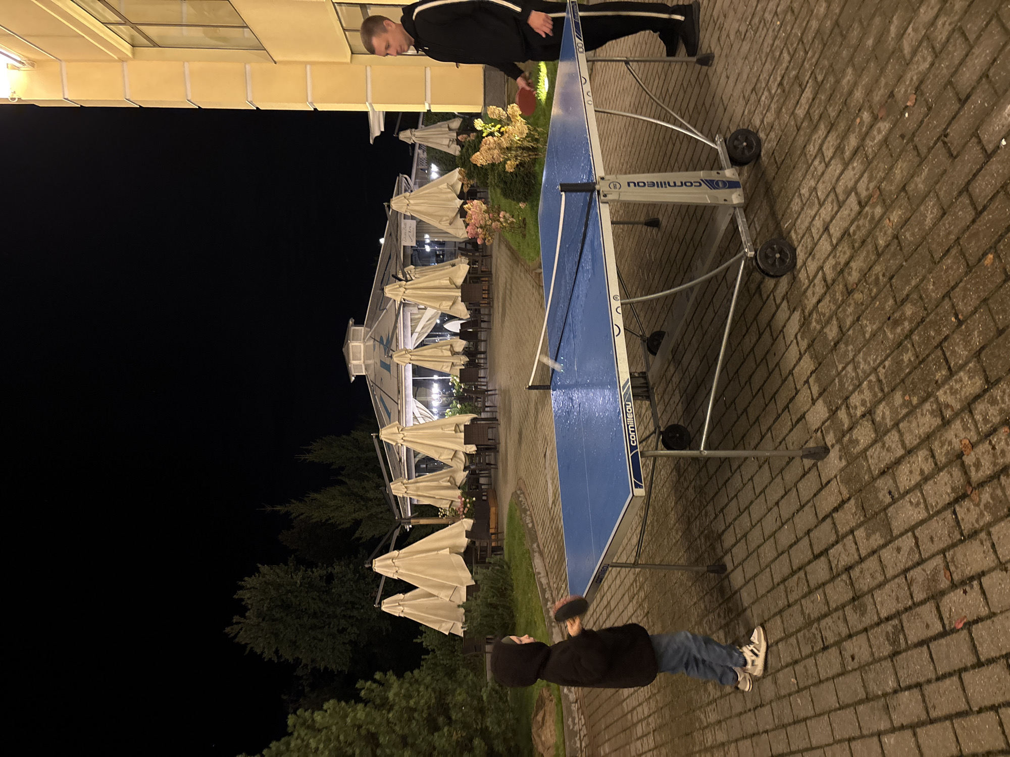 Two individuals are playing table tennis outdoors at night in an urban setting, likely in the city of Рождествено, Russia. The scene is illuminated by artificial lighting, casting a warm glow on the yellow building on the right and the outdoor patio area. The ground is paved with brick-like stones, which appear wet, suggesting recent rain or mist. A blue and silver table tennis table is positioned in the center of the frame. On the left, a child wearing a dark hooded jacket and jeans is holding a table tennis paddle, poised to strike. On the right, an adult male, dressed in a black tracksuit with white stripes, is also holding a paddle and facing the child across the table. In the background, several large, closed beige umbrellas are arranged around outdoor seating areas, and there are trees and bushes. The overall atmosphere is one of casual recreation.