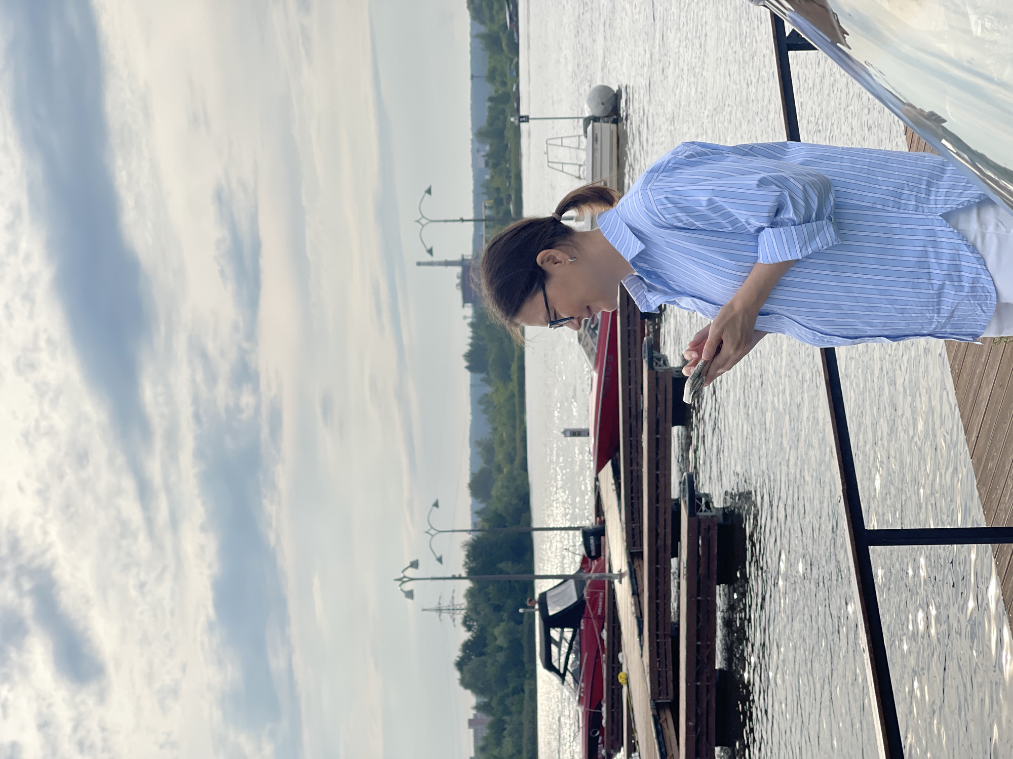 A woman is standing on a wooden dock, looking at her phone. She is wearing a blue and white striped shirt and white pants. She has brown hair pulled back in a ponytail and is wearing glasses. In the background, there are several boats docked in the water. The sky is cloudy. This image was taken in Izhevsk, Russia.