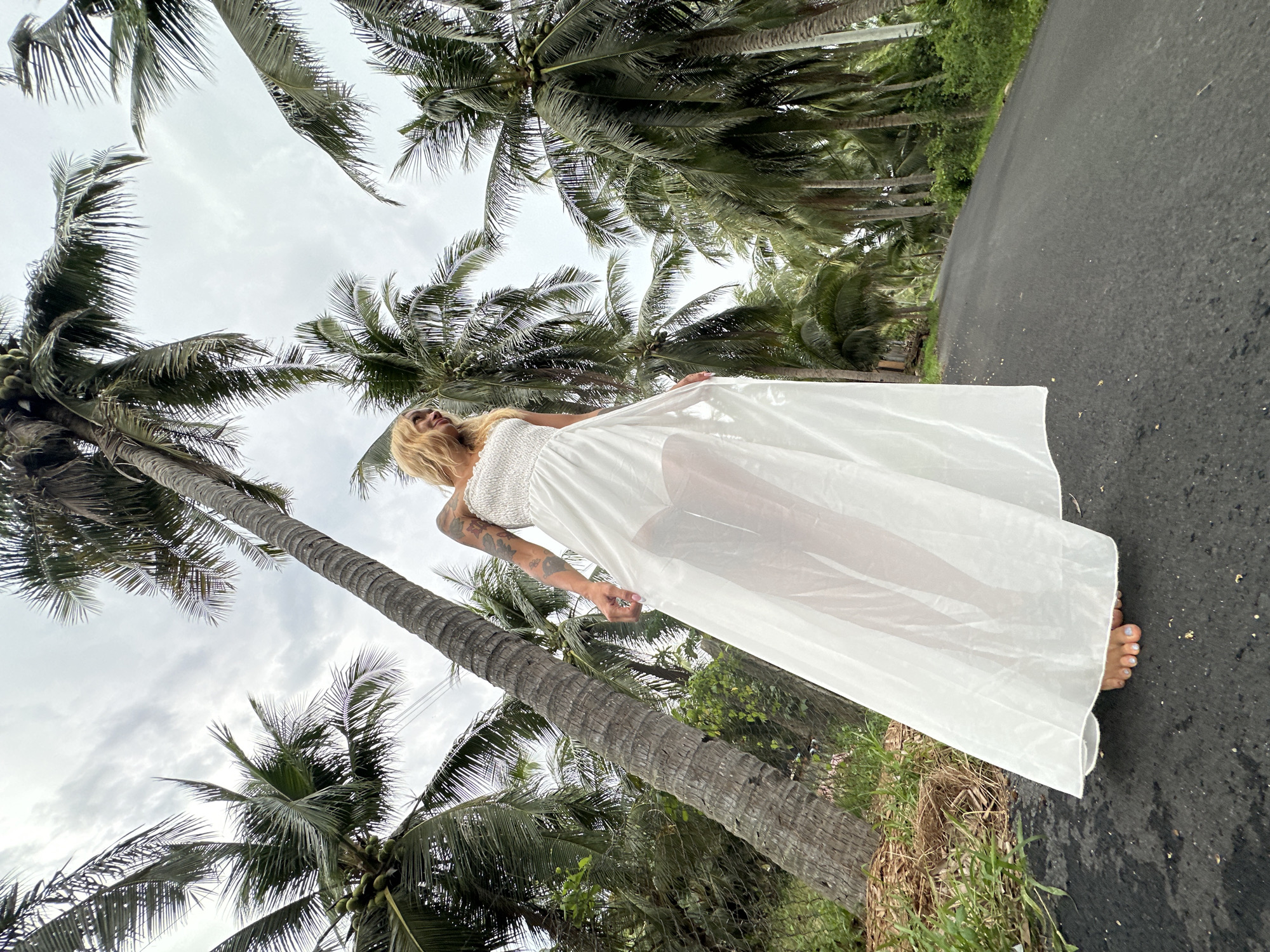 A woman wearing a white, sheer maxi dress walks on a paved road lined with tall coconut trees in Mũi Né, Vietnam. The woman, with blonde hair, stands barefoot on the dark asphalt, her dress flowing around her. She has tattoos on her arm. The road appears to be a quiet, rural route, possibly leading into the distance or towards a more populated area. The sky is overcast with clouds, suggesting a possibly humid or tropical climate.