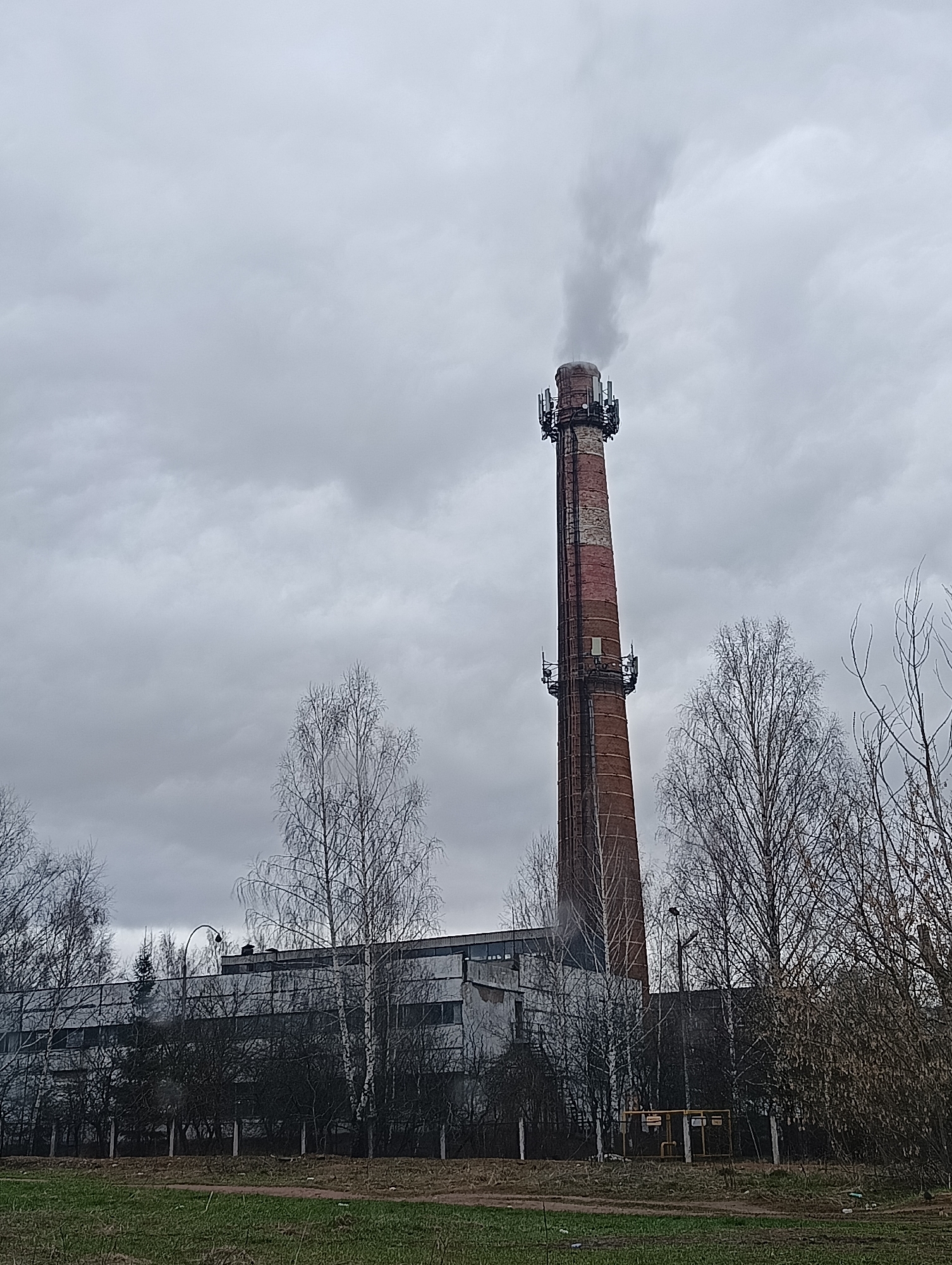 The image displays an industrial landscape under a heavily overcast, grey sky. Dominating the scene is a tall, slender red brick chimney, from which a plume of light grey smoke rises. The chimney features multiple metal platforms and antenna arrays attached at two different heights, indicating its dual use as an industrial stack and a telecommunications mast.
In the foreground and midground, a cluster of bare-branched deciduous trees, notably several slender birches, stands before a long, low industrial building with numerous dark windows. A street lamp post curves upwards near the building, partially obscured by trees. The ground in the immediate foreground is a mix of sparse green grass and dirt paths. To the right, a yellow metal structure, possibly part of a gas utility or industrial piping, is partially visible among the trees.
The bare trees suggest the season is late autumn, winter, or early spring. The overall impression is one of a functional industrial area on a cold or dreary day. No people or clear activities beyond the smoke emission are visible. Text on the yellow industrial structure is present but illegible due to distance and resolution.