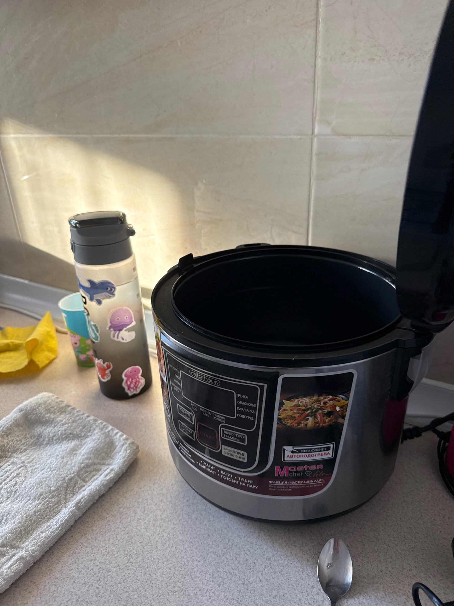 In a kitchen environment, likely within the city of Chișinău, Moldova, a metallic grey and red multi-cooker with its lid ajar is positioned on a speckled countertop. The multi-cooker features a control panel with various cooking programs labeled in Cyrillic script, including "ГРЕЧКА" (Buckwheat), "СУП/БОБОВЫЕ" (Soup/Legumes), "ПАР/ВАРКА" (Steam/Boiling), and "ПОДОГРЕВ" (Warming). A small image of paella is displayed on the panel. To the left of the multi-cooker sits a clear plastic water bottle adorned with stickers of marine life, including a dolphin and jellyfish. Next to the bottle is a blue and green plastic cup, also with stickers. A folded white terry cloth towel lies in the foreground to the left of the multi-cooker. A metal spoon is visible in the lower right portion of the frame. The background consists of a tiled wall with a light beige hue, illuminated by natural light casting shadows across the scene.