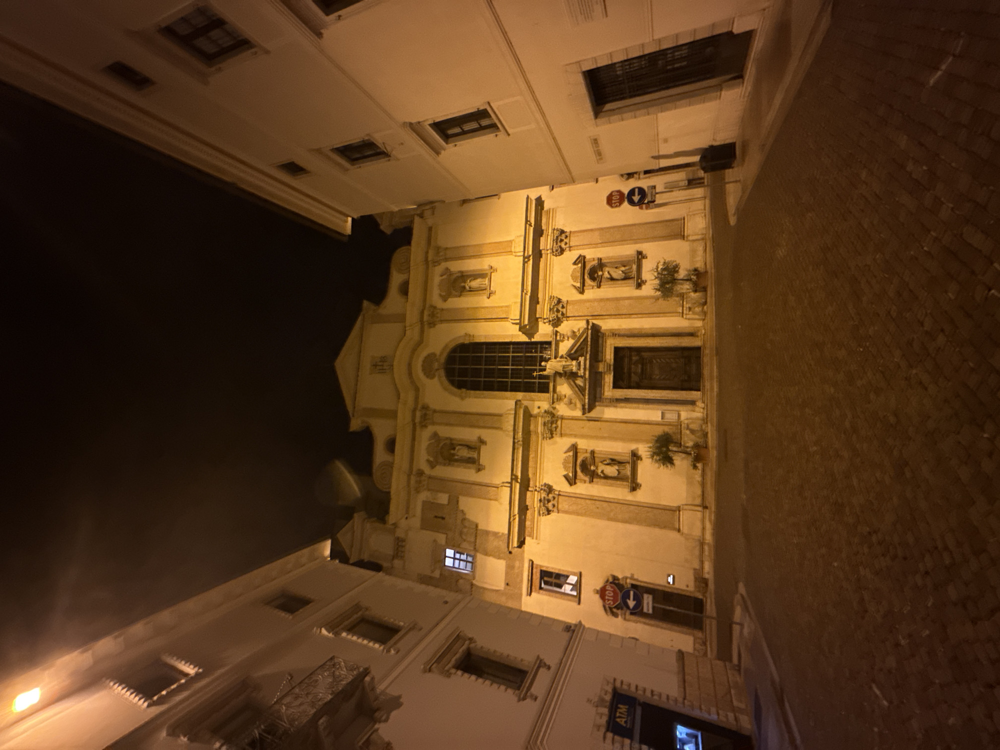 The image shows a wide shot of a street in Trento, Italy at night. A large, ornate building, possibly a church or a public building, dominates the center of the frame. It is illuminated by warm, artificial light, highlighting its architectural details such as columns, statues, and a large arched window. The street in front of the building is paved with cobblestones and is empty of people. Two buildings flank the central structure, one on the left with an ATM sign visible, and another on the right. Signs indicating "STOP" and a directional arrow are posted on the left side of the street. The overall atmosphere is quiet and still, with the nighttime illumination casting a strong glow on the scene.