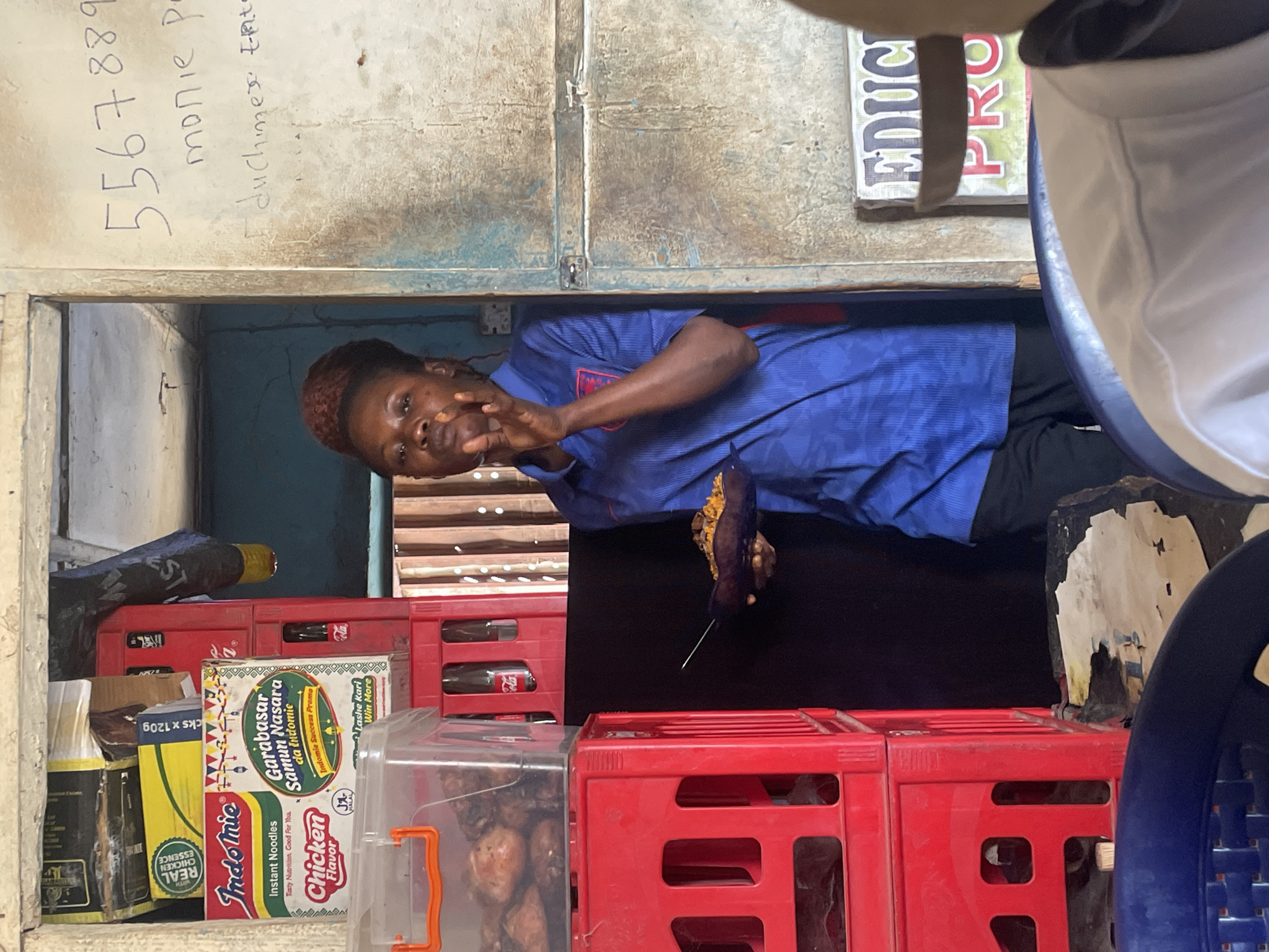 A woman stands in a doorway, possibly in Jos, Nigeria, holding a food item impaled on a stick. She is wearing a blue, patterned shirt. In front of her, red crates are stacked, displaying items like Indomie noodles and Coca-Cola bottles. A clear plastic container holds fried chicken. Handwritten text and phone numbers are on the wall above the doorway. A sign with the words "EDUC PRO" is partially visible on the right. A portion of a blue chair and a white object are in the foreground, obscuring parts of the scene.