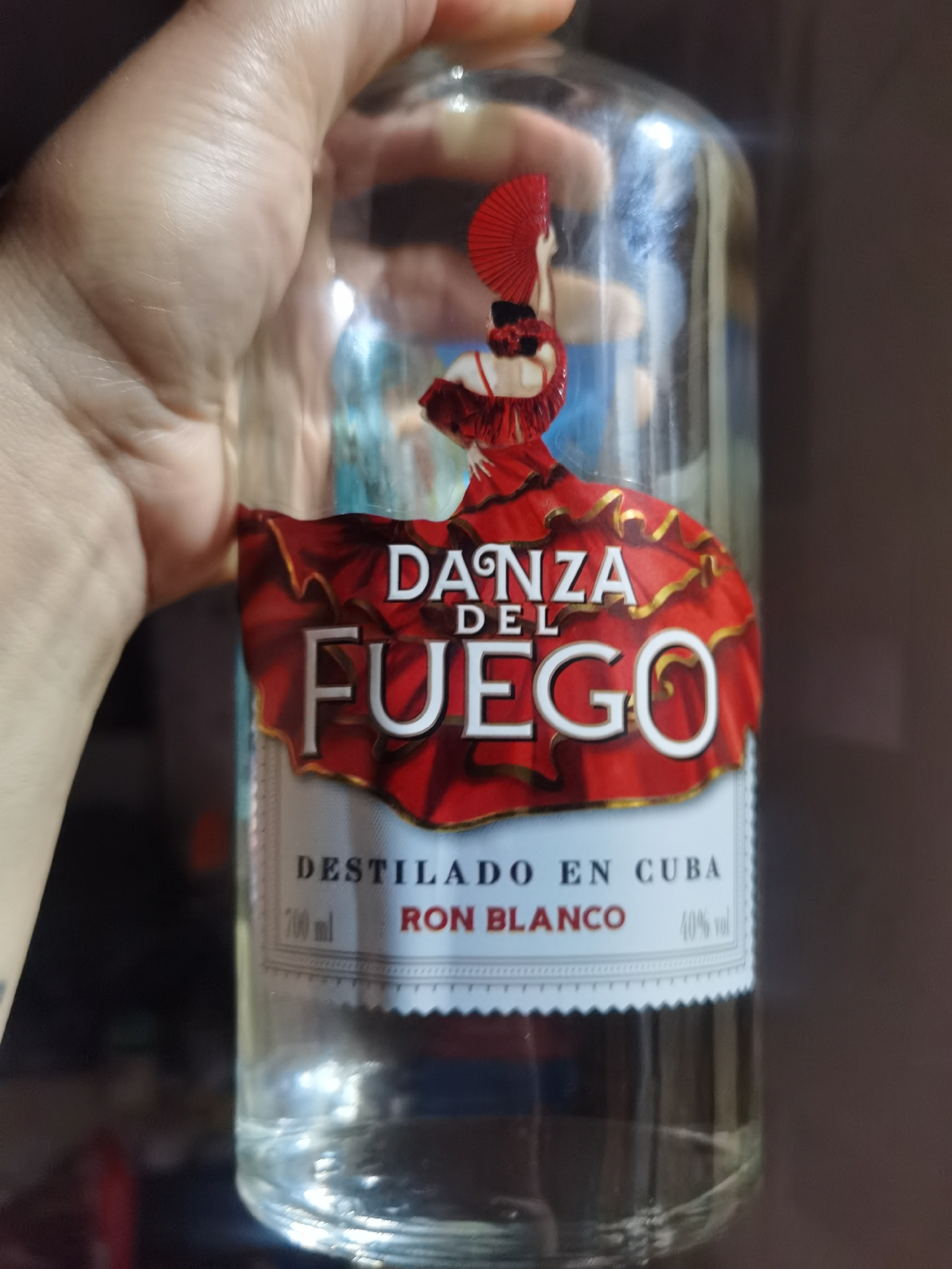 A close-up shot shows a clear glass bottle of "Danza del Fuego" rum, held by a person's hand. The label features a stylized image of a woman in a red dress with a fan and the words "Danza del Fuego," "Destilado en Cuba," and "Ron Blanco." The rum has 40% alcohol by volume and a net weight of 700 ml. The background is dark and blurred, obscuring the surrounding environment. There is no information in the image that would suggest a specific location in Chaikovsky, Russia.