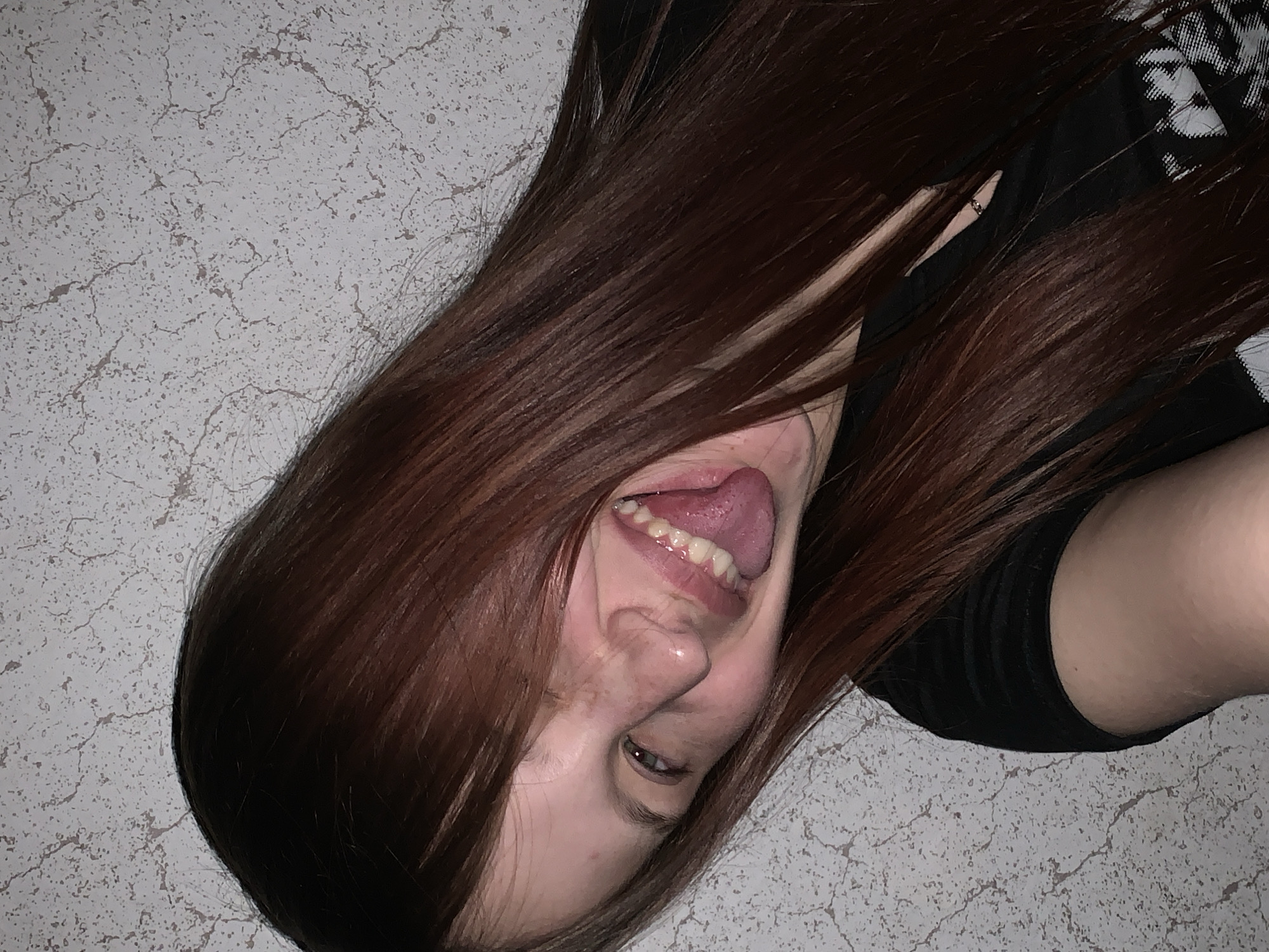 A young woman with long, straight brown hair is lying on her side against a grey speckled surface, possibly a floor or wall. She is sticking her tongue out and smiling, seemingly at the camera. She is wearing a black top. The location cannot be determined based solely on the visual information provided. The city of Фоки, Russia cannot be connected with the image without further information.