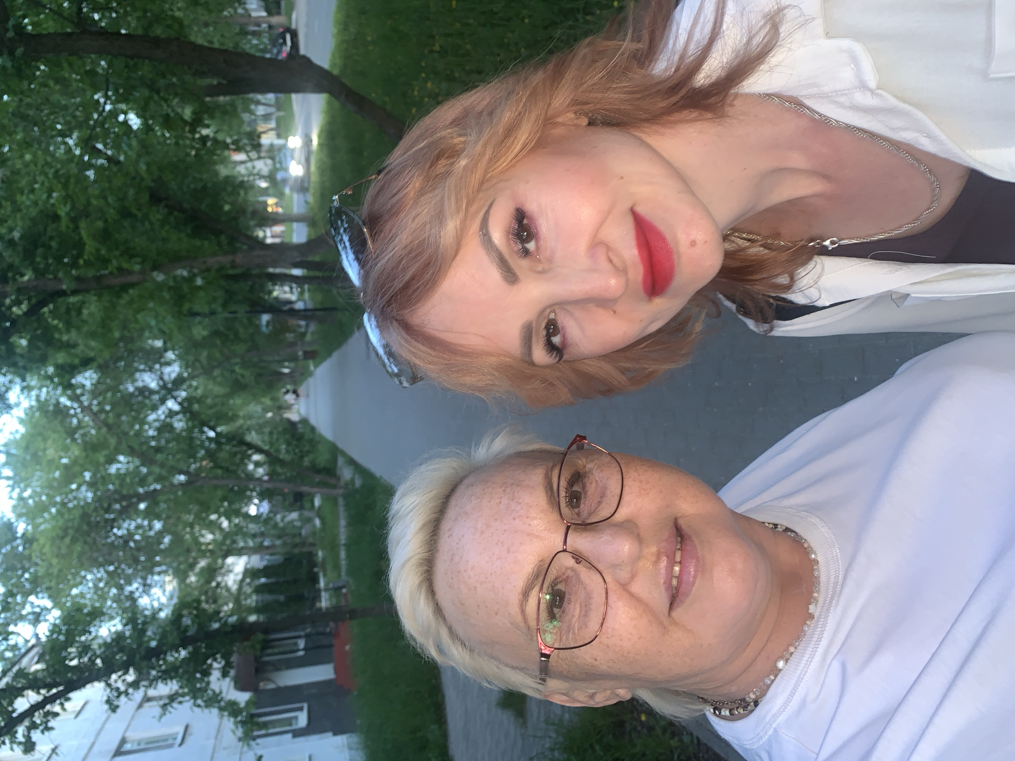 Two women are posing for a selfie outdoors. The woman on top has light brown hair, red lipstick, and sunglasses perched on her head. She is wearing a white jacket over a black top and a silver necklace. The woman below has short blonde hair and is wearing eyeglasses and a white t-shirt. She also has a beaded necklace. They are standing on a sidewalk with a green grassy area and trees in the background. In the distance, there's a building and streetlights, suggesting a residential area in the city of Chaikovsky, Russia.