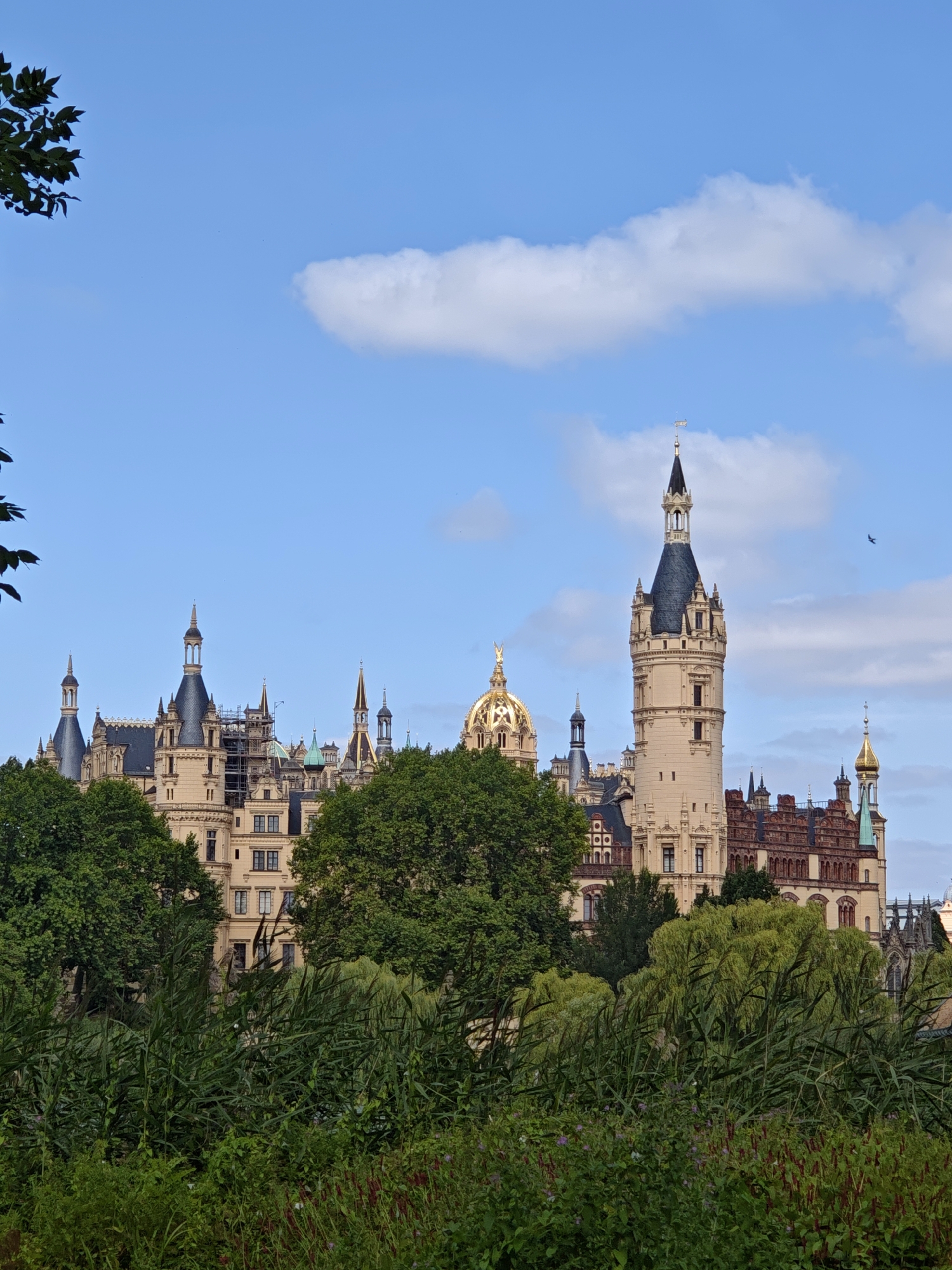 This image captures a view of Schwerin Castle (Schweriner Schloss), a prominent landmark located on an island in the city of Schwerin, Germany. The castle, a grand architectural complex, is situated in the background, partially obscured by lush green trees and vegetation in the foreground. The castle itself features numerous turrets, spires, and ornate details, with a large central tower being particularly noticeable. The sky above is a clear blue, dotted with a few fluffy white clouds. A small bird is visible flying in the upper right portion of the sky. The scene appears to be a daytime shot, with natural lighting illuminating the castle and its surroundings.